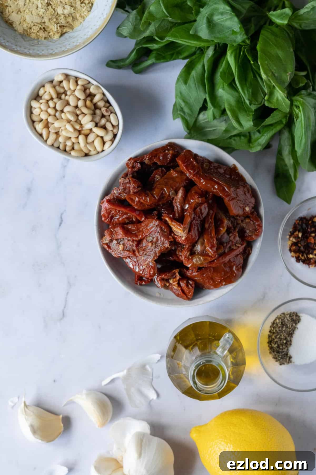 Vegan Sun-Dried Tomato Pesto Ingredients - Fresh Basil, Sun-Dried Tomatoes, Pine Nuts, Garlic, Lemon, Olive Oil for Dairy-Free Pesto Rosso. #veganingredients #plantbasedcooking A beautiful flat lay of fresh ingredients laid out on a marble surface, ready to be transformed into pesto. It includes a jar of sun-dried tomatoes, fresh basil, pine nuts, garlic cloves, a lemon, and a bottle of olive oil.