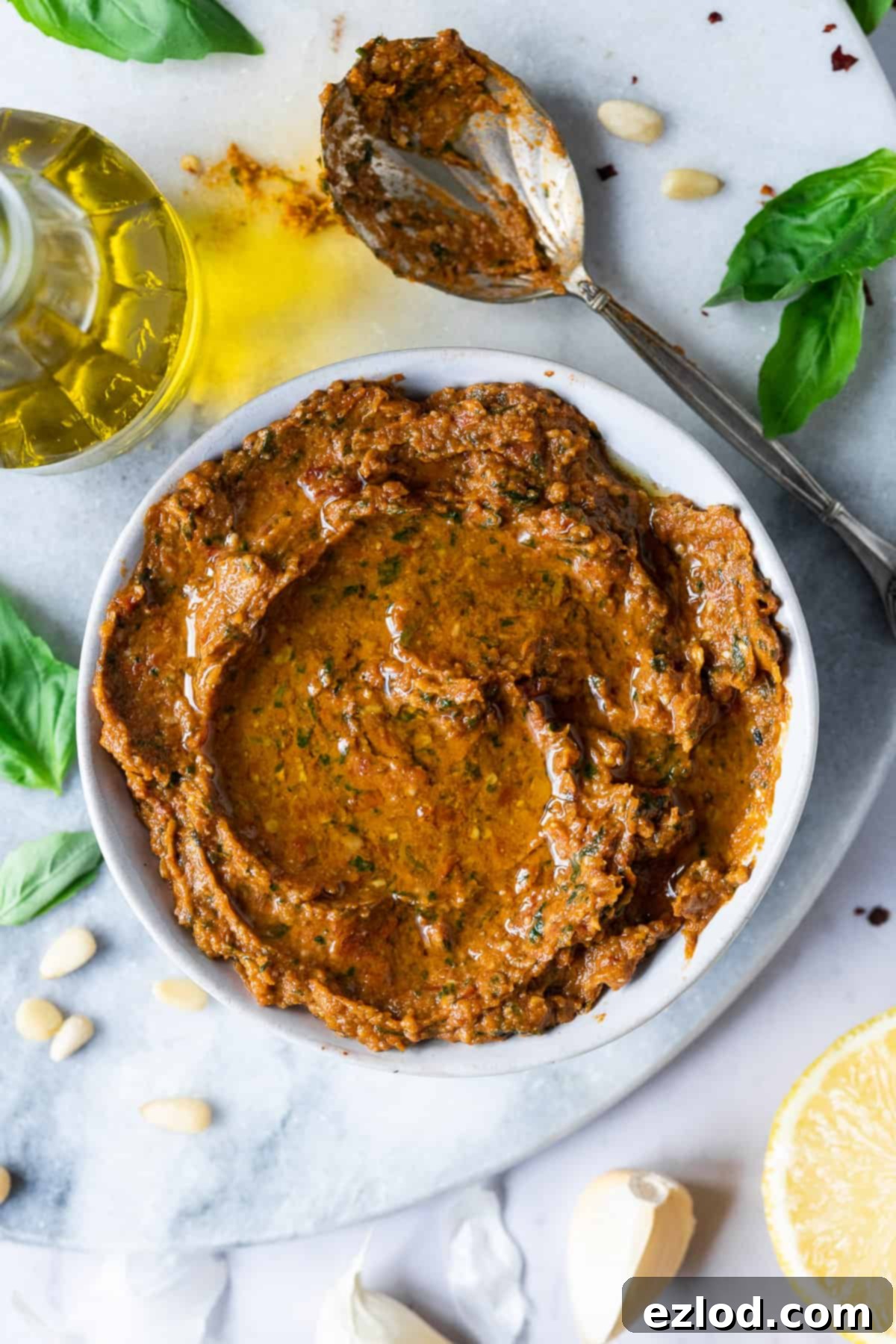 Freshly Made Vegan Sun-Dried Tomato Pesto - Dairy-Free Pesto Rosso Bowl with Ingredients. Easy and Delicious Plant-Based Condiment. #homemadepesto #pestorecipe A vibrant bowl of homemade red pesto sits elegantly on a marble board, accompanied by a wooden spoon, fresh basil leaves, a bottle of olive oil, whole garlic cloves, and a bright yellow lemon, showcasing the fresh components.