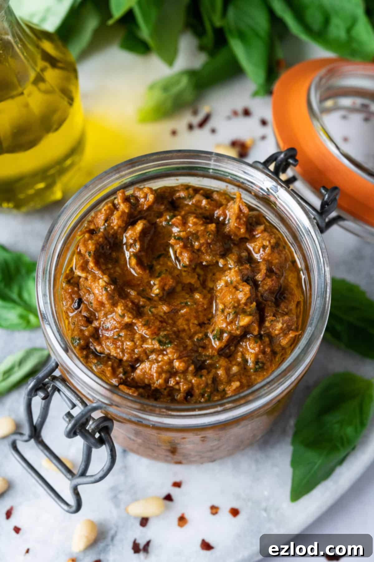 Jar of Vegan Sun-Dried Tomato Pesto - Homemade Dairy-Free Pesto Rosso with Fresh Basil. Perfect for Storing and Enjoying Later. #veganfood #pestoideas A close-up view of a glass jar filled with luscious vegan red pesto, elegantly placed on a marble surface and adorned with scattered fresh basil leaves, emphasizing its natural freshness.