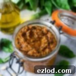 Vibrant Vegan Sun-Dried Tomato Pesto 3 A jar of vibrant red vegan sun-dried tomato pesto on a marble board with fresh green basil leaves and a clear bottle of olive oil.