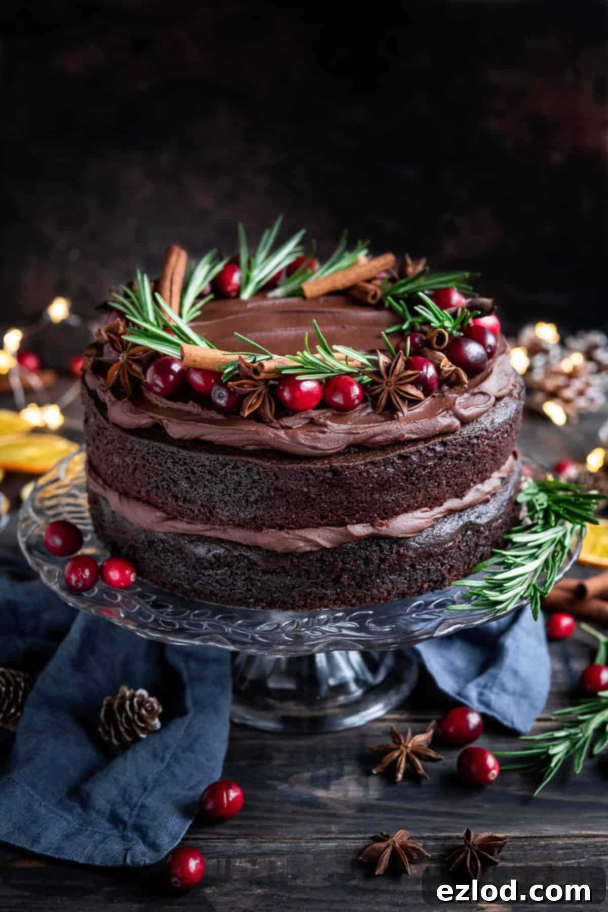 Festive Vegan Mulled Wine Chocolate Cake Vegan mulled wine chocolate cake topped with cranberries, rosemary, star anise and cinnamon sticks on a glass cake stand on a wooden table.