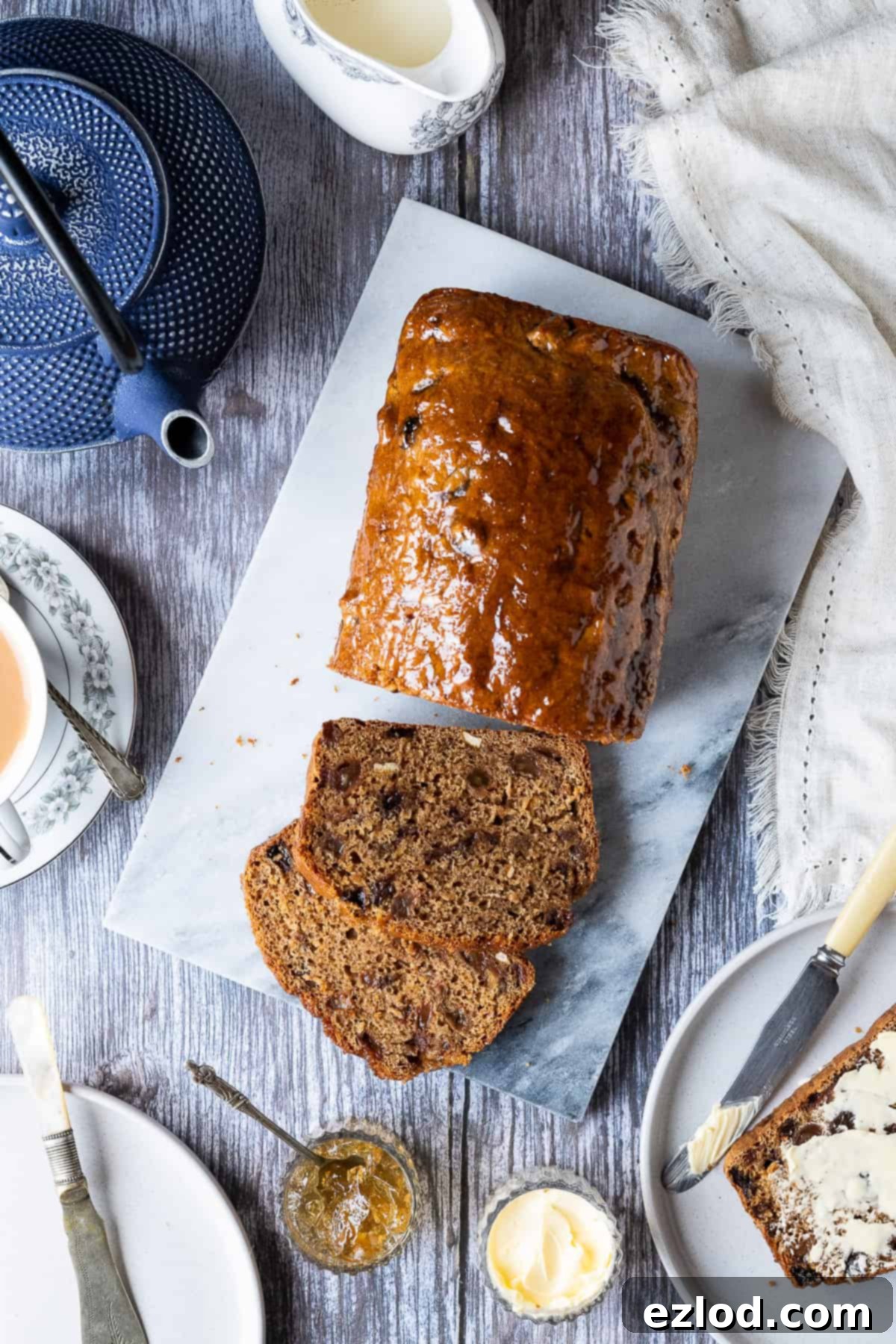 Homemade Vegan Earl Grey Tea Loaf: Easy, fat-free, and fruit-filled. Perfect for an eggless and dairy-free snack. #veganbaking #vegancake #eggless #loafcake Vegan Earl Grey tea loaf displayed on a marble board on a wooden table, surrounded by a blue teapot, matching plates, knives, bowls of vegan butter and marmalade, a cup of tea, and a jug of plant-based milk – an inviting scene for afternoon tea.