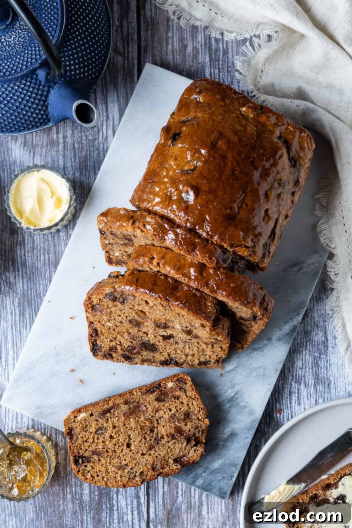 Vegan Earl Grey Tea Loaf Slices – easy, fat-free, fruity, eggless, and dairy-free for British tea time. #veganbaking #vegancake #eggless #loafcake Top down shot of a sliced vegan tea loaf on a marble board set on a wooden table, alongside a blue teapot, inviting a cozy tea time.