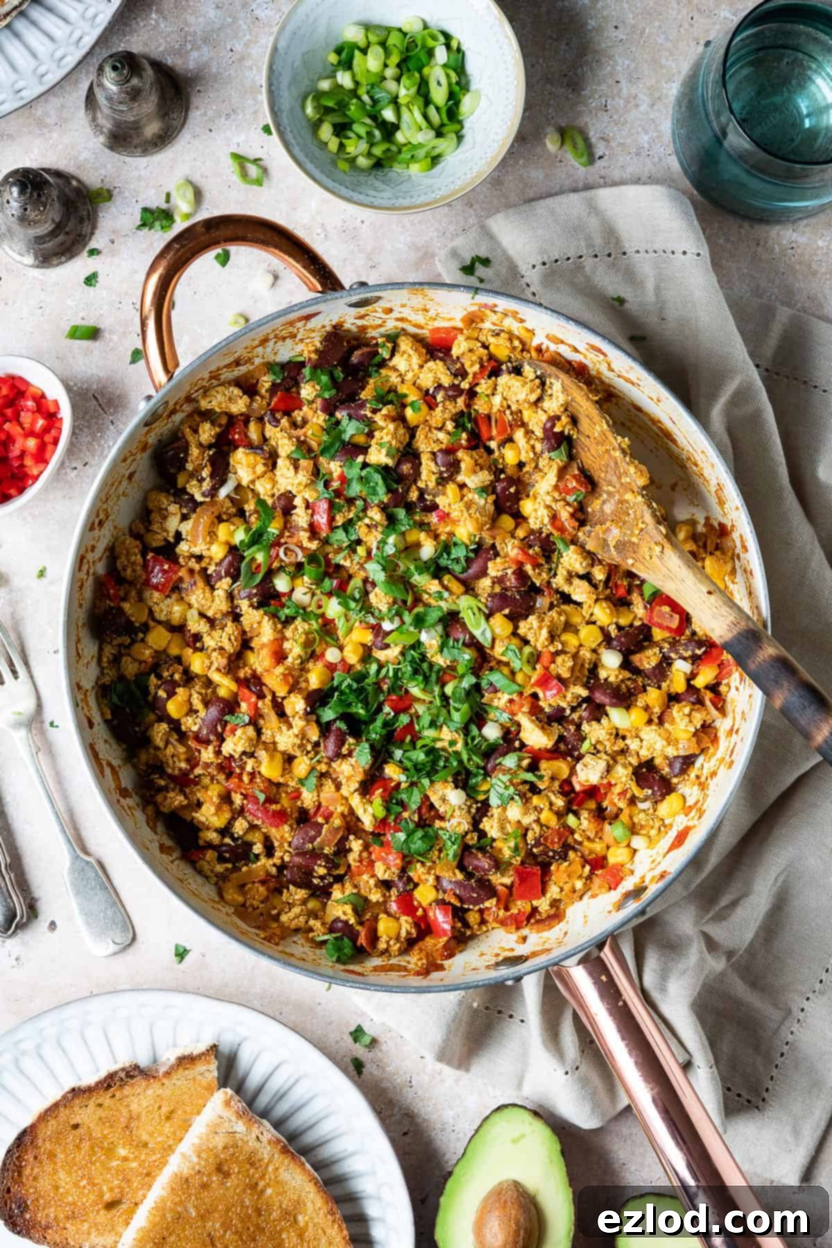 Mexican Style Tofu Scramble - Easy Vegan Protein-Packed Meal for Breakfast, Lunch, or Dinner with Fresh Veggies and Authentic Flavors #veganbreakfast #veganmeal #plantbased A large white frying pan holds a generous portion of Mexican style tofu scramble, garnished with fresh spring onions and coriander. It is artfully arranged on a light wooden surface, surrounded by two plates of golden-brown toast, a perfectly halved avocado, a rustic beige cloth, a refreshing glass of water, and small bowls of vibrant chopped spring onion and red chilli, suggesting fresh garnishes for the meal. The scene is bright and inviting, highlighting a healthy and flavorful vegan breakfast or brunch.
