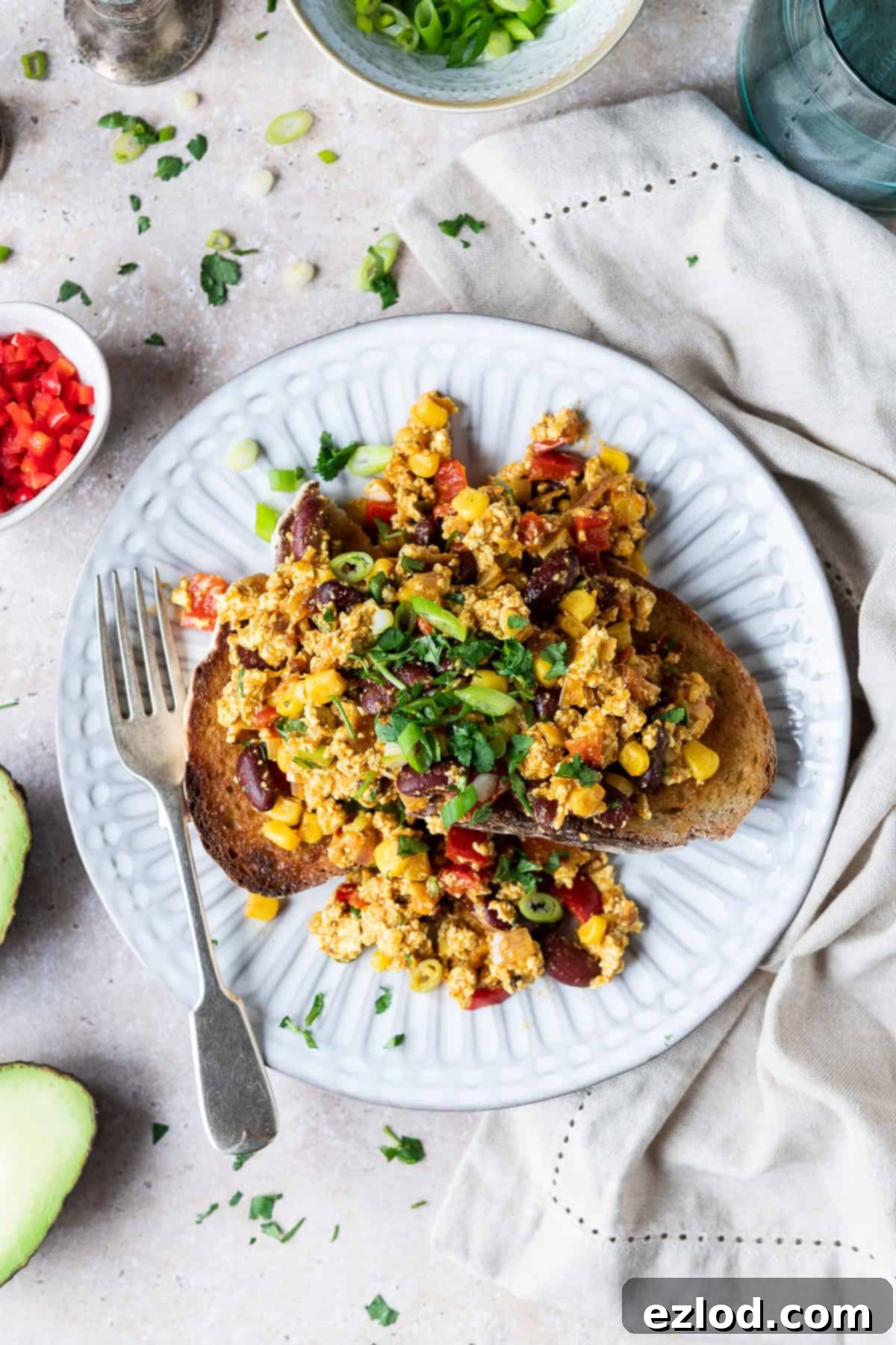 Mexican Style Tofu Scramble on Toast with Avocado and Fresh Herbs - Delicious Vegan Breakfast, Lunch, or Dinner #plantbasedrecipe #veganfood A beautifully composed flat lay showcases a white plate with Mexican style tofu scramble served on toast, accompanied by a silver fork. The setting is enhanced with a scattered garnish of fresh green spring onions and vibrant coriander leaves. Surrounding the plate are a perfectly halved avocado, a clear glass of water, a soft beige cloth, and small white bowls holding chopped red chilli and additional spring onions, ready for customization. This inviting scene captures the essence of a wholesome, flavorful, and easy vegan meal.