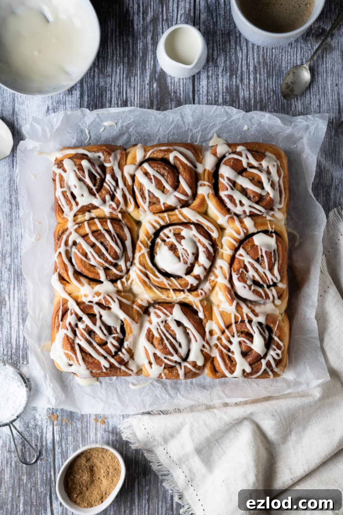 Vegan cinnamon rolls - these delicious cinnamon rolls are made using the tangzhong method which makes them incredibly soft, fluffy, squishy and moreish. Eggless and dairy-free. #veganbread #veganbaking #eggless #dairyfree Vegan cinnamon rolls artfully arranged on baking parchment on a wooden table, surrounded by a bowl of glaze, a jug of non-dairy milk, a cup of coffee, and a bowl of cinnamon.