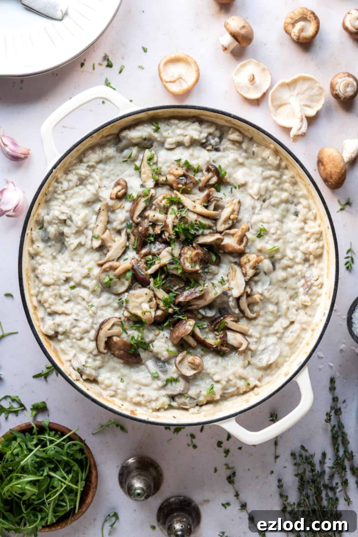 Vegan mushroom risotto with truffle oil – this creamy vegan risotto is full of flavour thanks to earthy mushrooms and truffle oil. Topped with garlic butter shiitake mushrooms it is truly a meal to impress! #veganmeal #plantbased #dairyfree A large pan of creamy vegan mushroom risotto surrounded by fresh ingredients: raw mushrooms, thyme sprigs, salt and pepper shakers, a bowl of fresh rocket, garlic skins, and an empty plate.