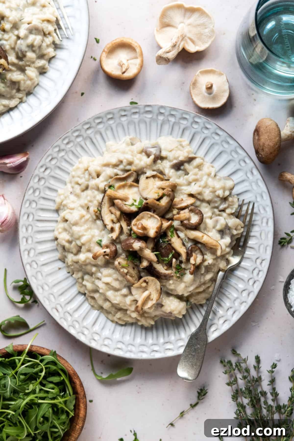 Vegan mushroom risotto with truffle oil – this creamy vegan risotto is full of flavour thanks to earthy mushrooms and truffle oil. Topped with garlic butter shiitake mushrooms it is truly a meal to impress! #veganmeal #plantbased #dairyfree A full plate of vegan mushroom risotto, generously topped with garlic shiitake mushrooms, surrounded by a fresh rocket salad, thyme, raw garlic cloves, a glass of water, and more fresh mushrooms.