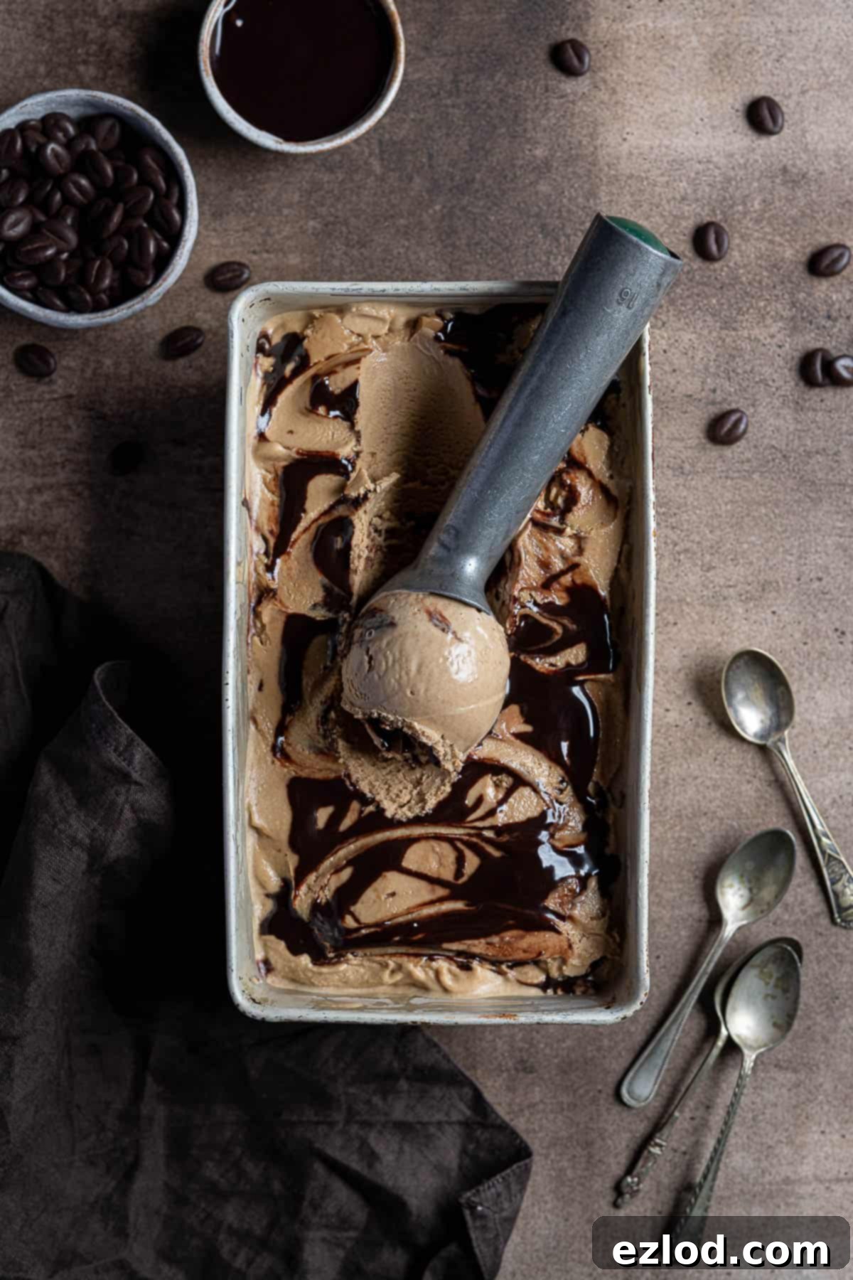 Serve up this incredible vegan coffee ice cream, super smooth and creamy, with a delightful coffee flavor and a rich chocolate fudge swirl. Easy to make and completely coconut-free, with oats as the secret ingredient! Vegan coffee ice cream in a metal tin on a brown surface with a bowl of chocolate sauce, bowl of chocolate coffee beans, a brown cloth and metal tea spoons.