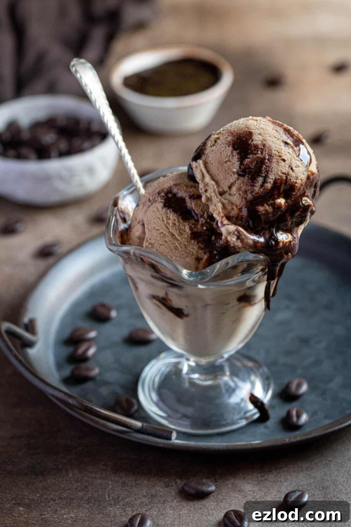 Enjoy two delicious scoops of our homemade vegan coffee ice cream, a smooth and creamy treat with a rich coffee flavor and chocolate fudge swirl, made without coconut. Two scoops of vegan coffee ice cream in a glass on a metal tray.
