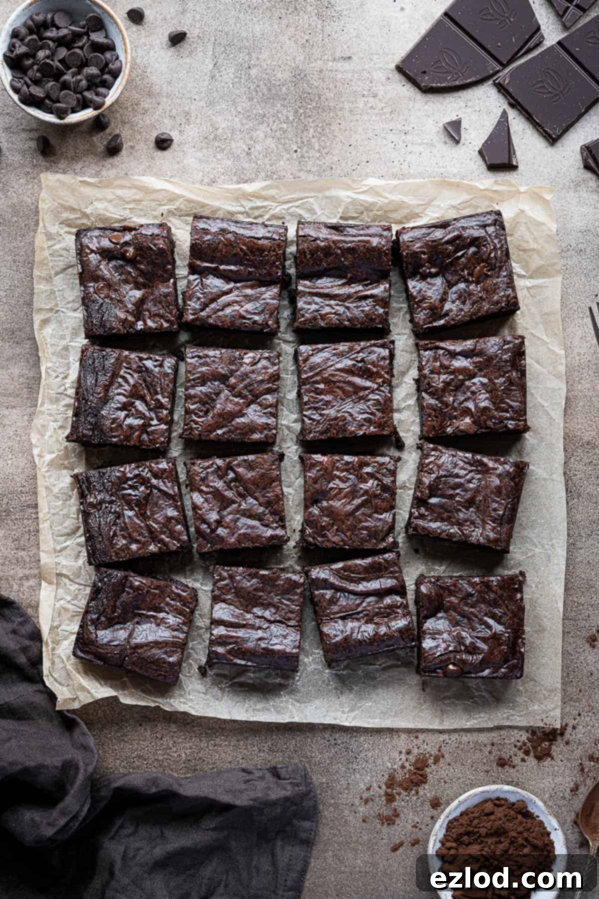 Seriously Fudgy Plant-Powered Brownies 3 Fudgy vegan brownies on baking parchment, surrounded by bowls of chocolate chips, cocoa powder, a broken dark chocolate bar, and a brown cloth.