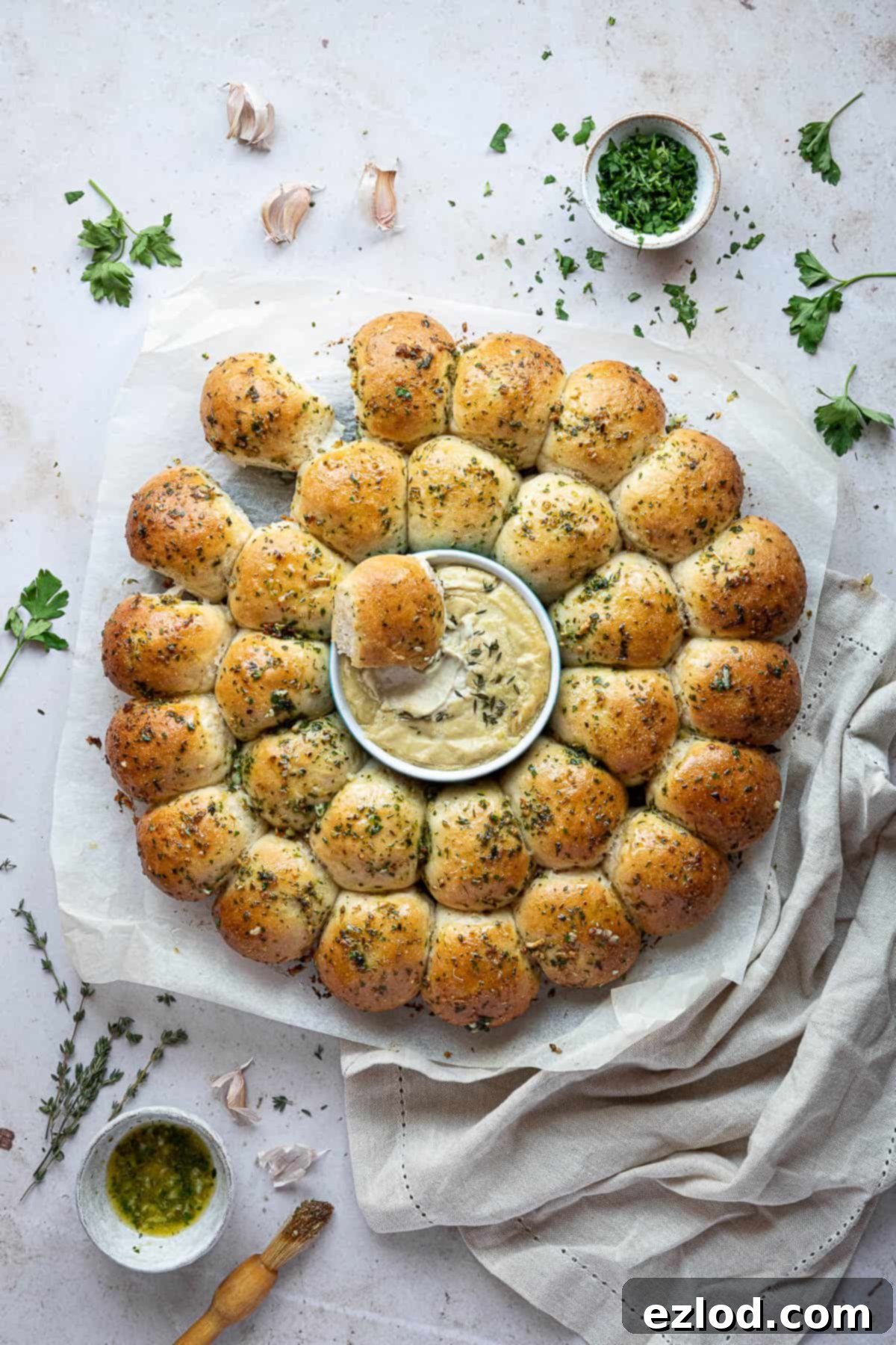 Pillowy Vegan Garlic Dough Balls 14 A partially torn apart vegan garlic dough ball wreath on baking parchment, with bowls of garlic butter and herbs visible nearby, inviting diners to enjoy.