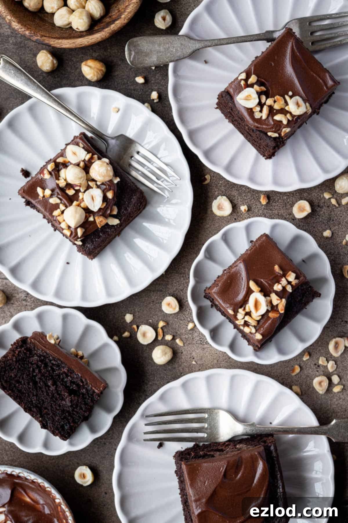 Decadent Vegan Chocolate Hazelnut Cake 3 Perfectly cut squares of moist vegan chocolate hazelnut cake, generously frosted with a rich ganache and garnished with chopped hazelnuts, are presented on elegant white plates, ready to be enjoyed.