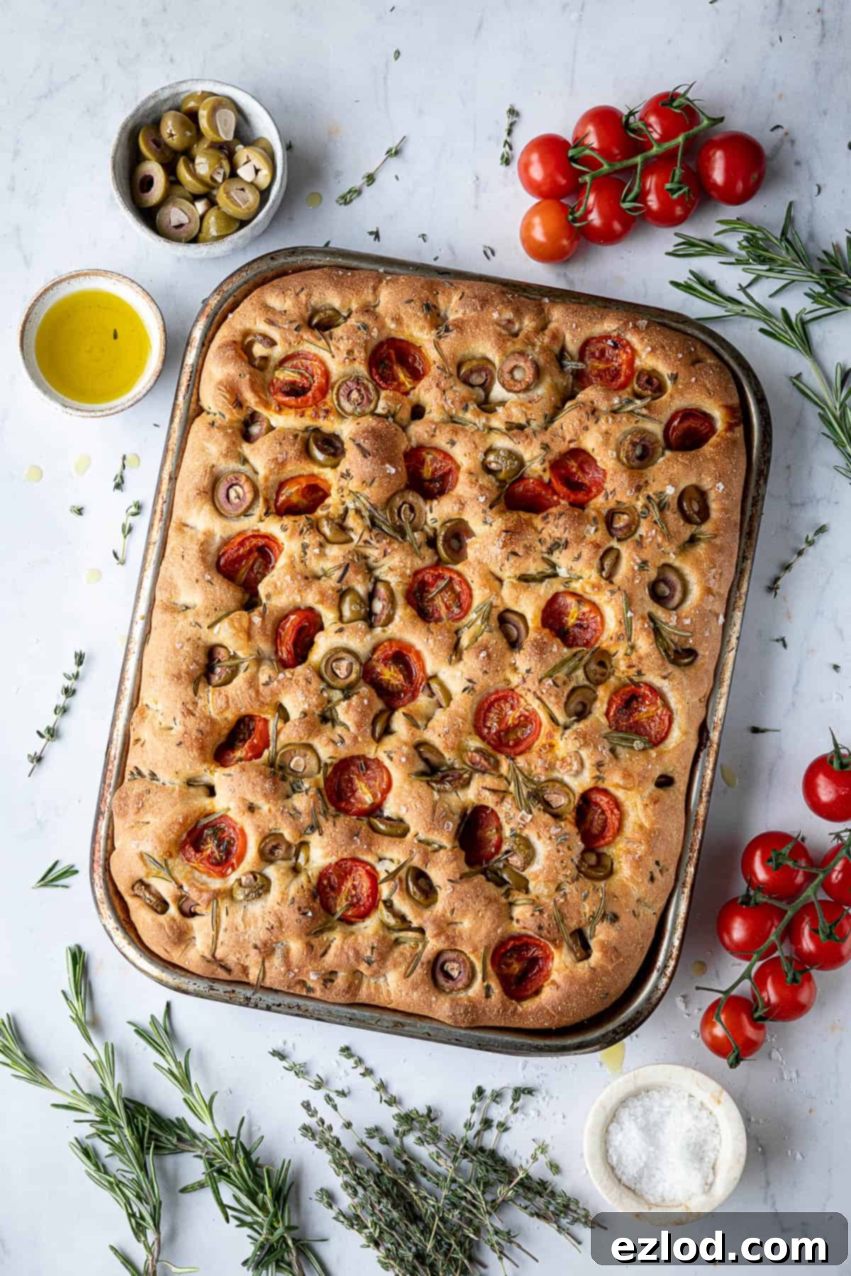 Baking Bliss and BBC Maestro Insights 2 Tomato olive focaccia surrounded by herbs, tomatoes and bowls of olives, salt and olive oil, highlighting its fresh ingredients and inviting aroma.