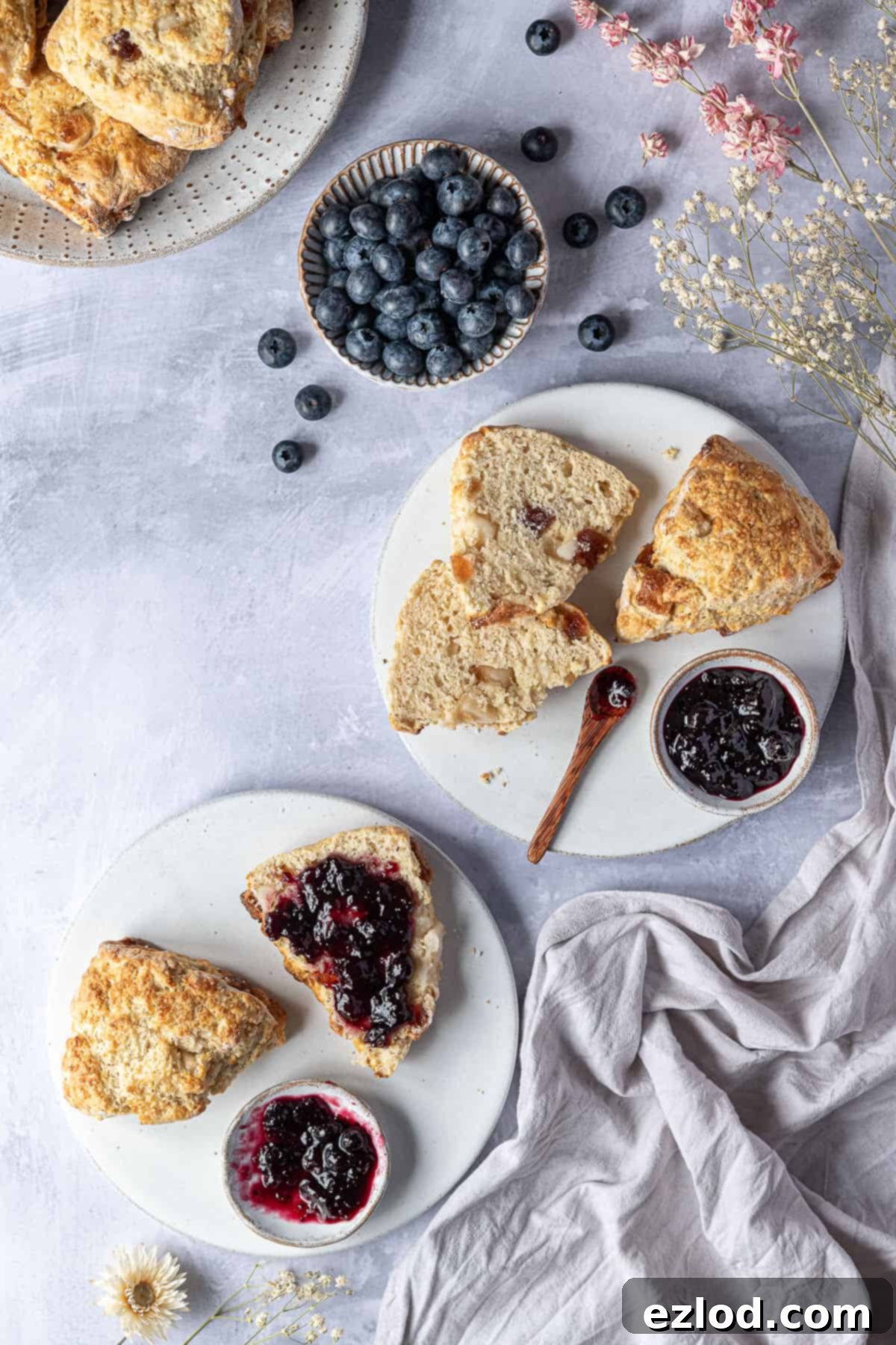 Light, Moist, and Easy Vegan Marzipan Cherry Scones - Perfect for Any Occasion! Three white plates of vegan marzipan cherry scones with bowls of jam, a bowl of blueberries, a grey cloth and dried flowers, highlighting the perfect texture.