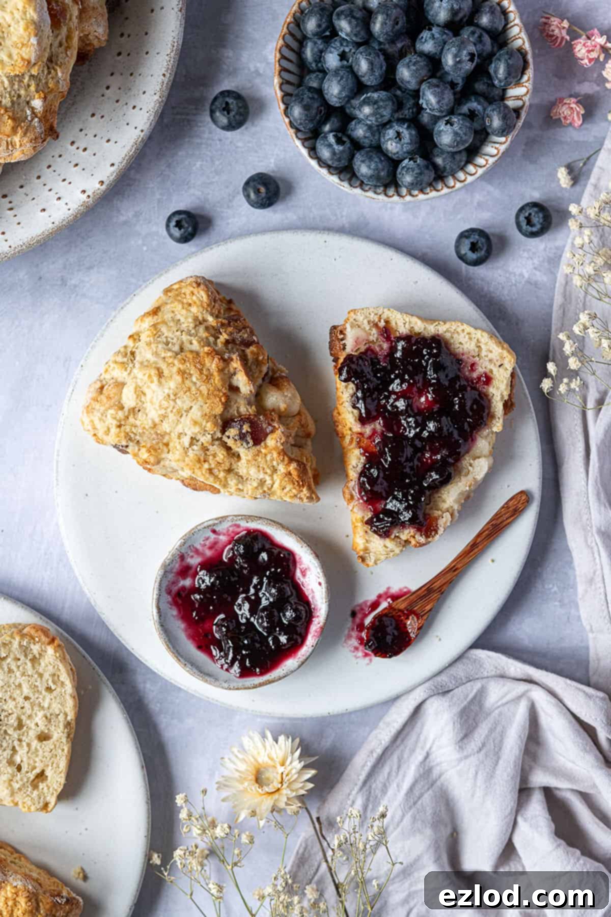 Serve Your Vegan Marzipan Cherry Scones with Jam and Fresh Berries! A beautifully presented vegan marzipan cherry scone, sliced in half with jam, surrounded by a bowl of blueberries, dried flowers, grey linen, and plates of additional scones for a vibrant tea spread.