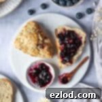 English Title Crafting 5 A delicious vegan marzipan cherry scone sliced in half with jam, surrounded by a bowl of blueberries, dried flowers, grey linen, and plates of scones.