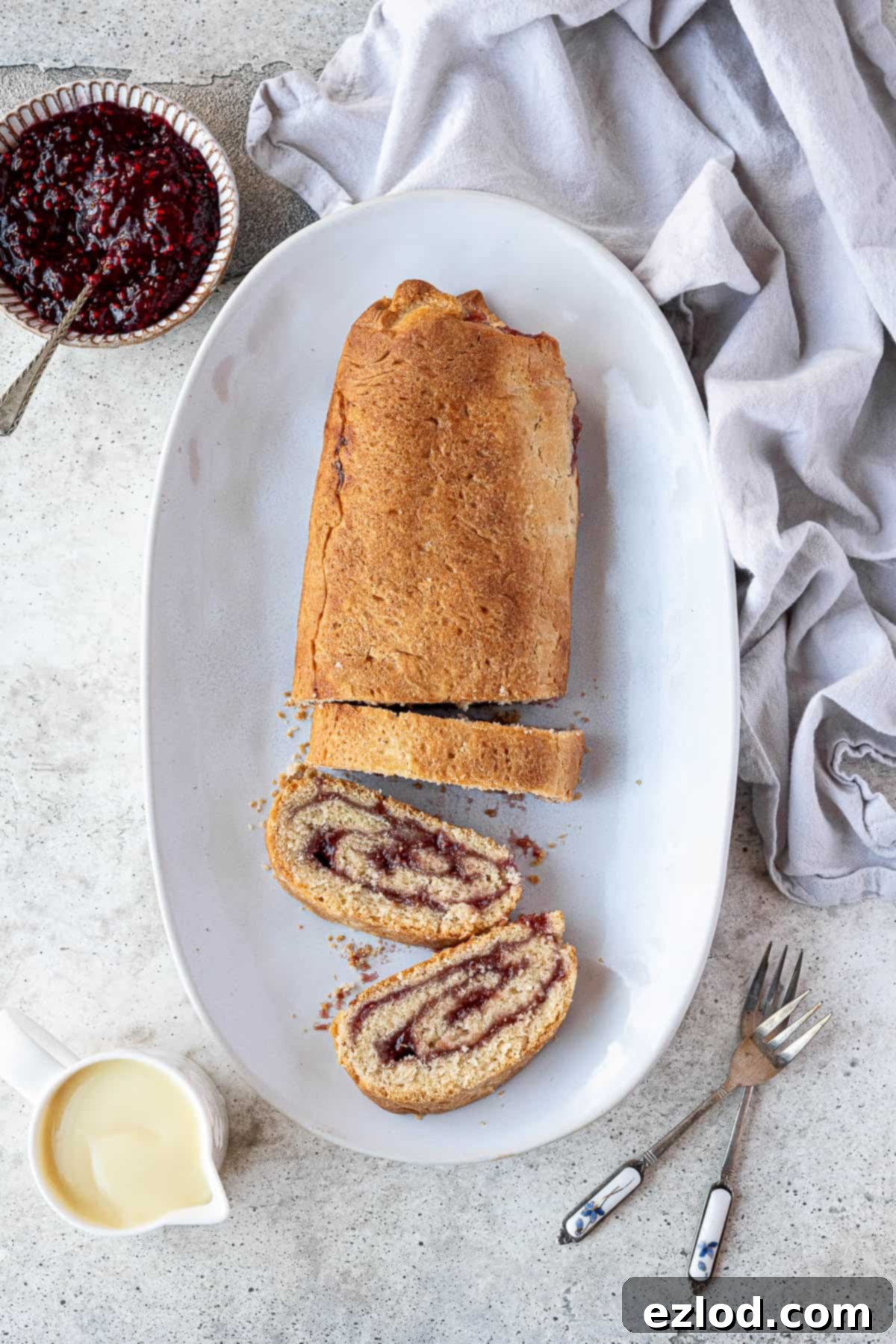 Luscious Vegan Jam Roly-Poly 14 A beautifully presented vegan jam roly-poly on a white plate, accompanied by a small jug of vegan custard and a bowl of red jam.