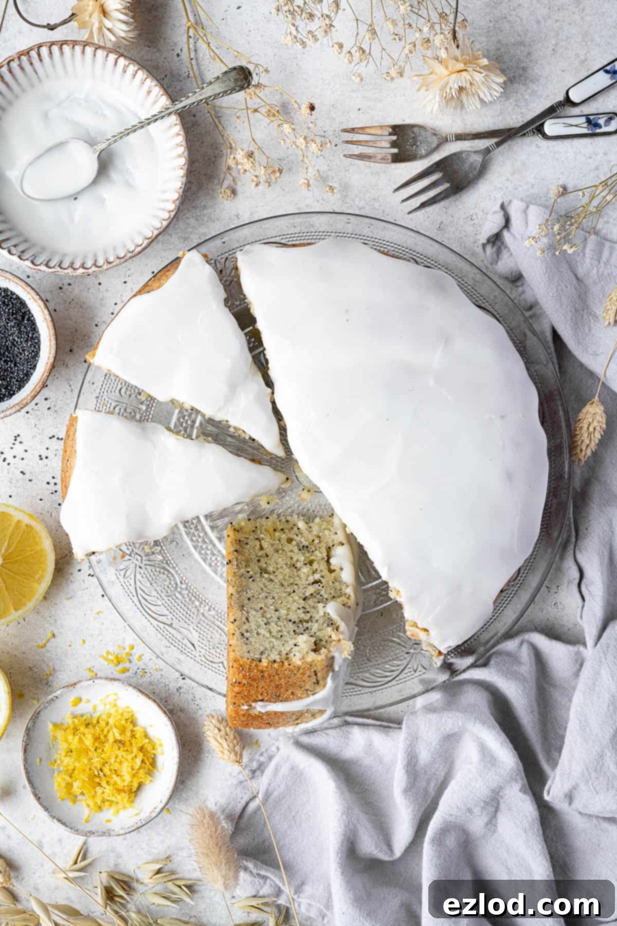 Zesty Vegan Lemon Poppy Seed Cake 3 A whole vegan lemon poppy seed cake presented on a glass plate, surrounded by small bowls containing fresh lemon zest, poppy seeds, and a vibrant lemon icing, illustrating the key ingredients.