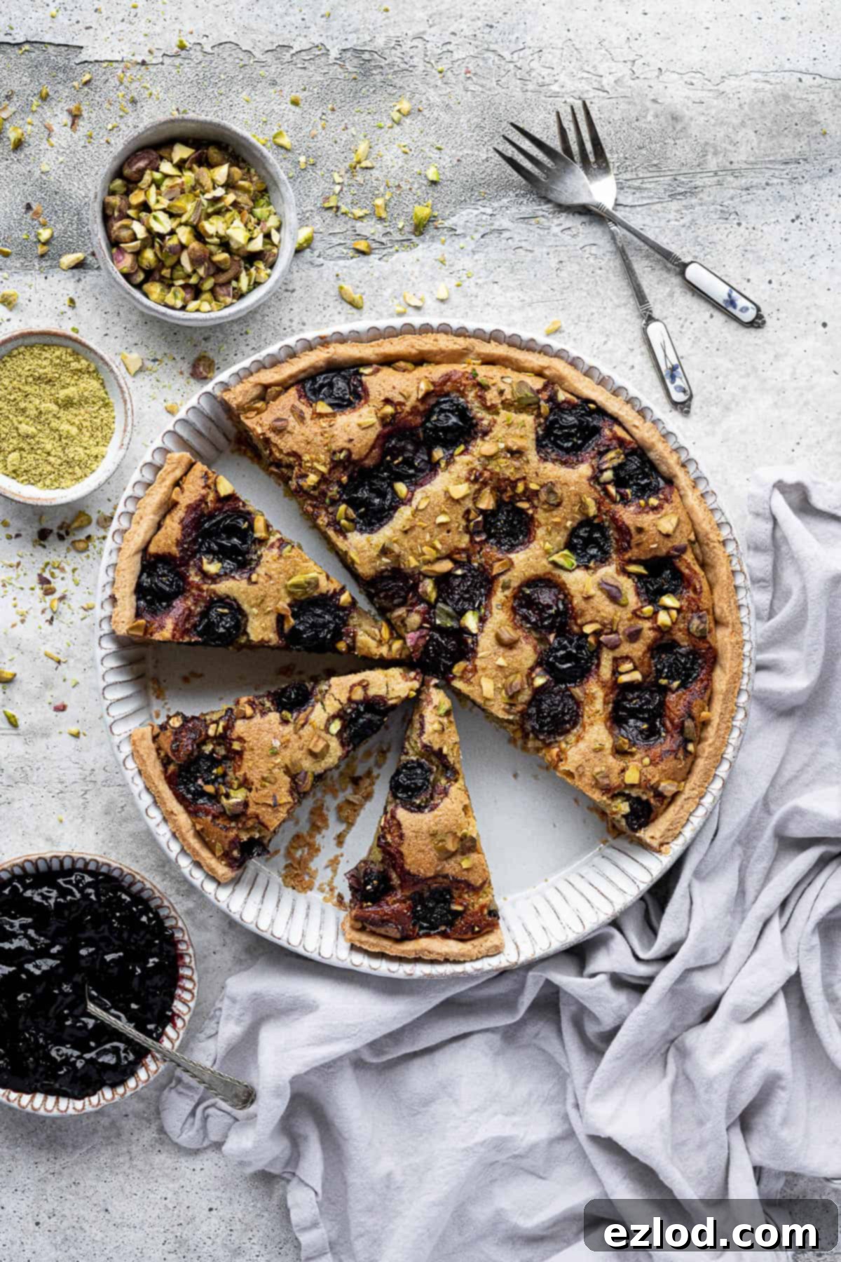 Heavenly Vegan Pistachio Cherry Frangipane Tart 11 The sliced pistachio cherry frangipane tart on a grey surface with bowls of jam and pistachios.