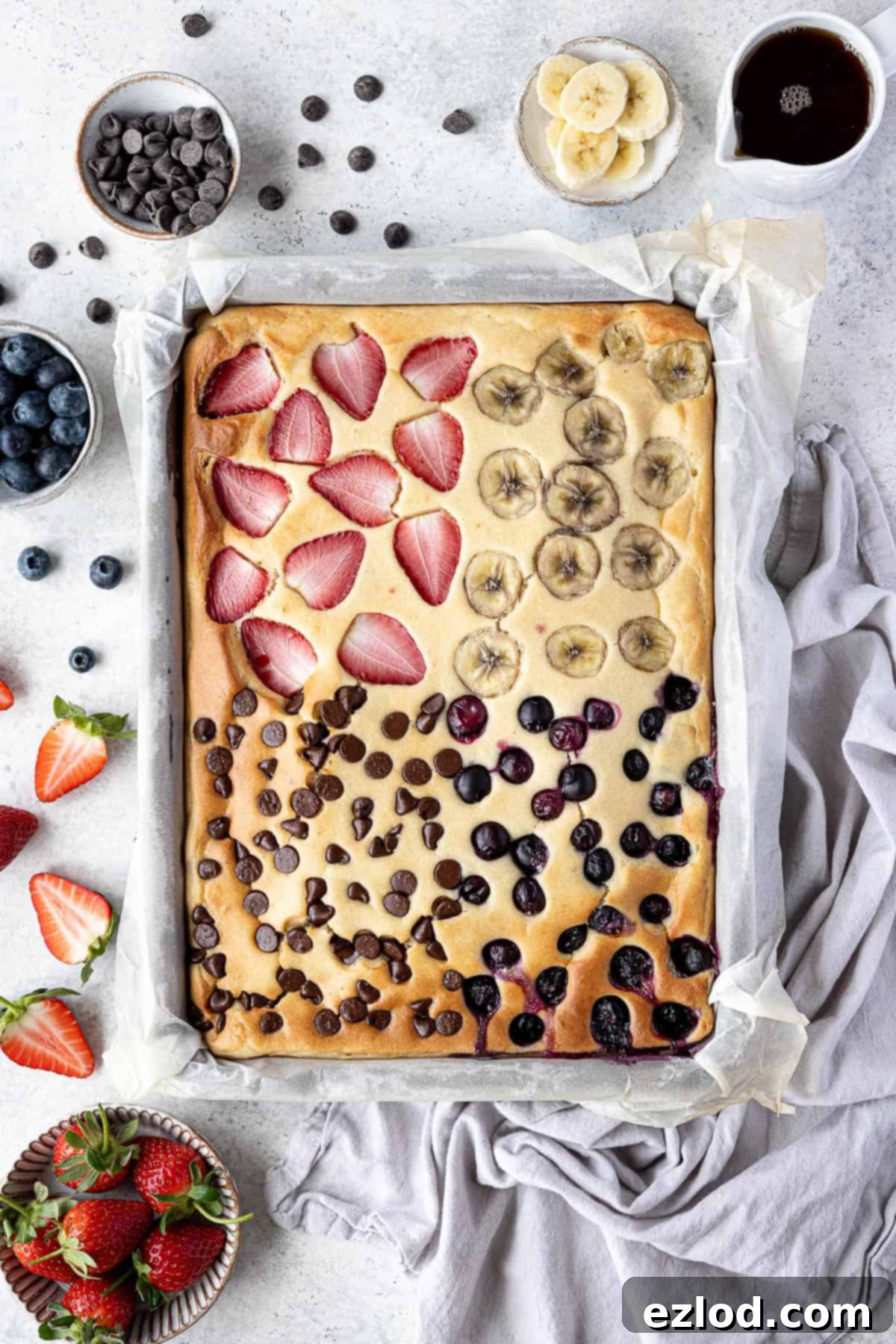 Effortless Vegan Sheet Pan Pancakes 2 Vegan sheet pan pancakes with bowls of chocolate chips, strawberries, blueberries, banana and a jug of maple syrup.
