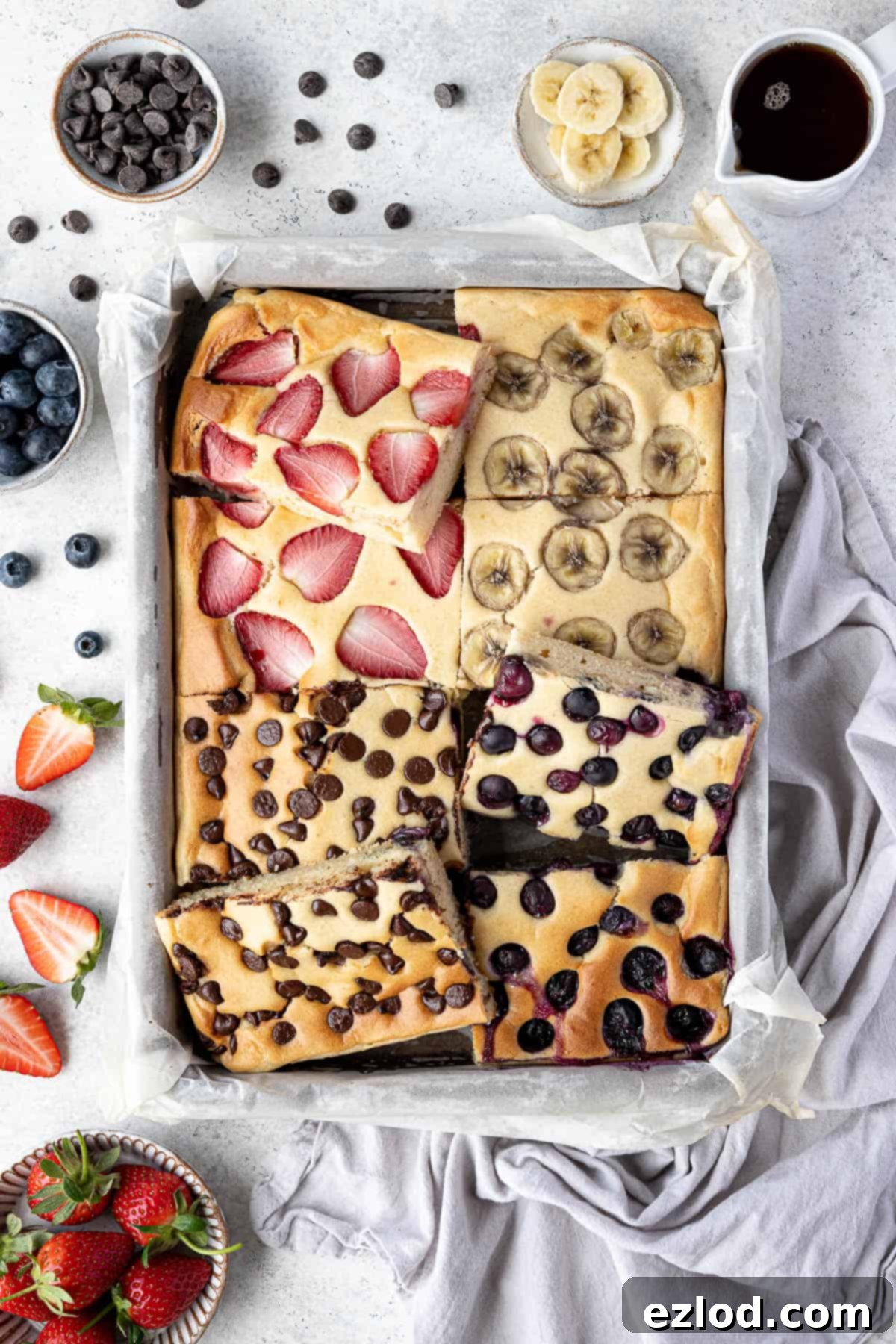 Effortless Vegan Sheet Pan Pancakes 9 Sliced vegan sheet pan pancakes in a baking tray with bowls of chocolate chips, strawberries, blueberries, banana and a jug of maple syrup.