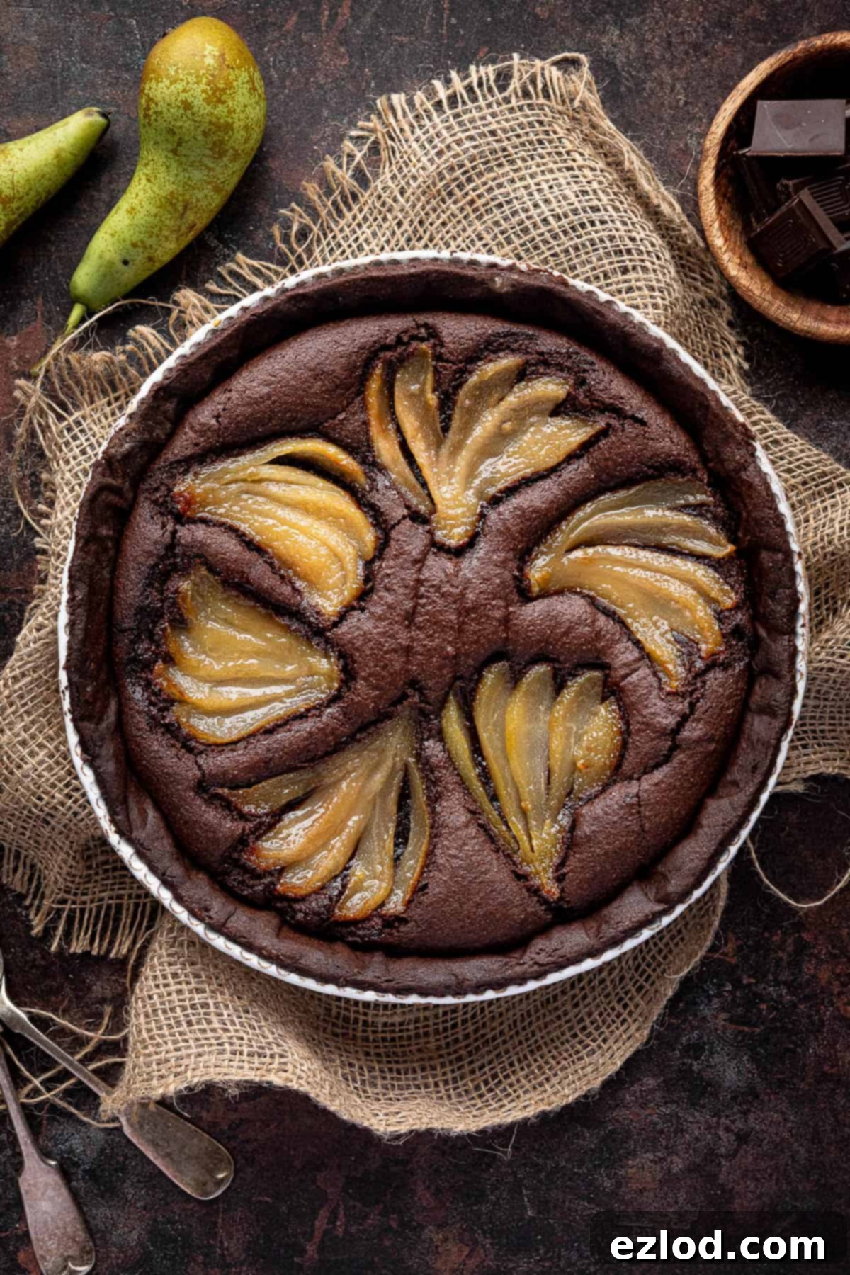 Rich Vegan Chocolate Pear Almond Tart 2 A stunning slice of vegan chocolate pear frangipane tart on a dark surface, surrounded by whole pears, a bowl of melted chocolate, and elegant forks.