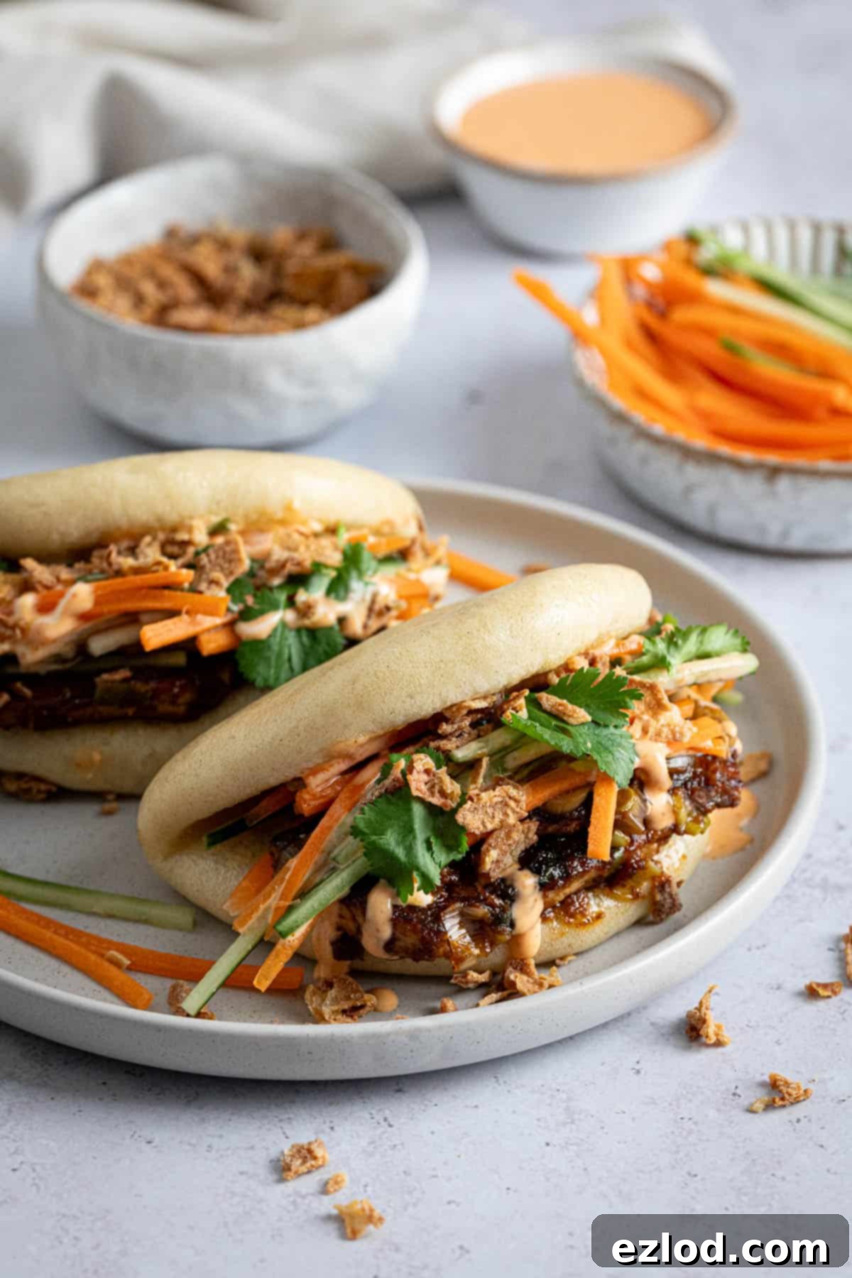 Pillowy Vegan Hoisin Tofu Bao Buns 4 Two hoisin tofu bao buns on a plate, with small bowls of shredded vegetables and sriracha mayo, ready to be assembled.