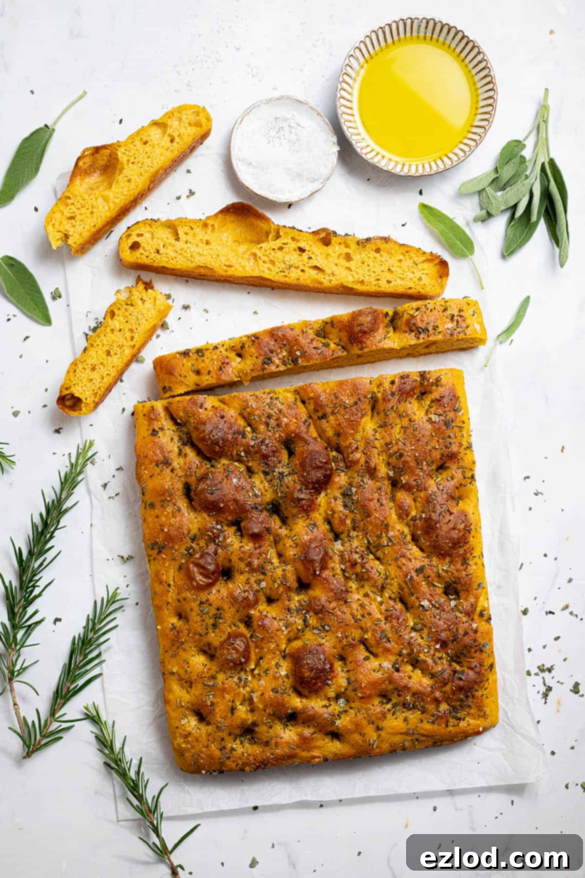 Autumn Spiced Vegan Pumpkin Focaccia 13 Pumpkin focaccia on a white surface with fresh herbs and bowls of olive oil and sea salt.