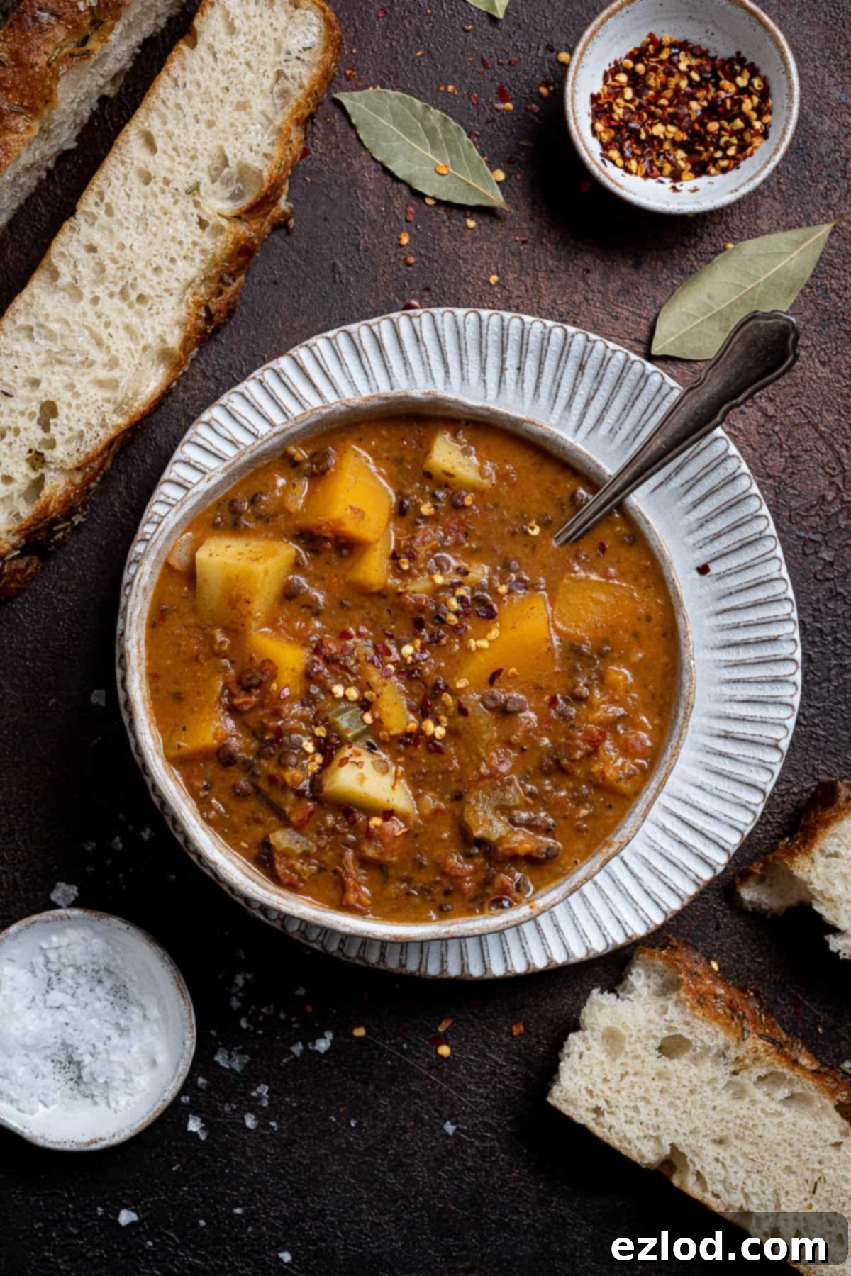 Mediterranean Sunshine Lentil Soup (Vegan) 5 A bowl of sun dried tomato lentil soup on a white plate with slices of crusty bread, alongside small bowls of salt and chilli flakes for extra seasoning, on a wooden surface.