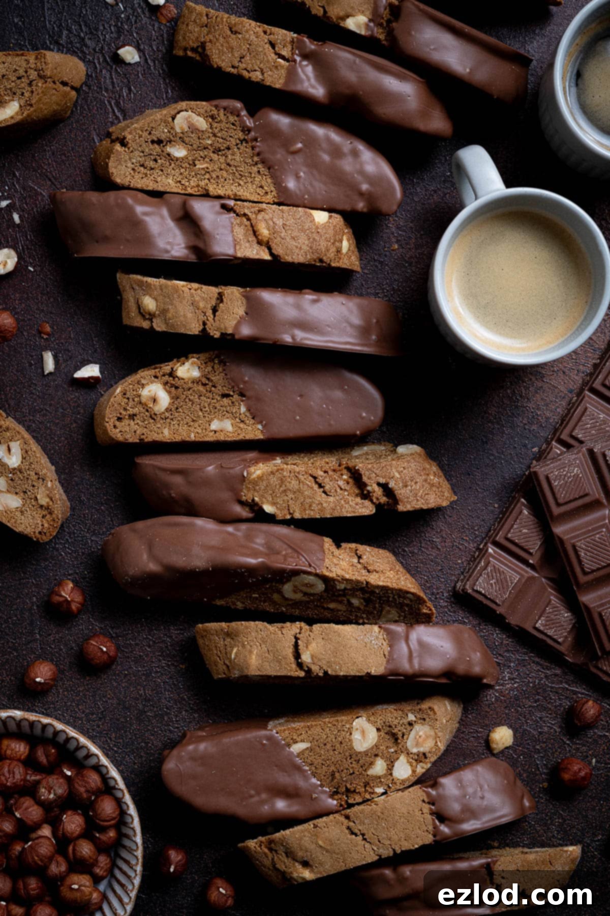 Plant-Based Coffee Hazelnut Biscotti 11 Vegan coffee hazelnut biscotti arranged in a row on a dark surface with cups of espresso, a bar of chocolate and a bowl of hazelnuts.