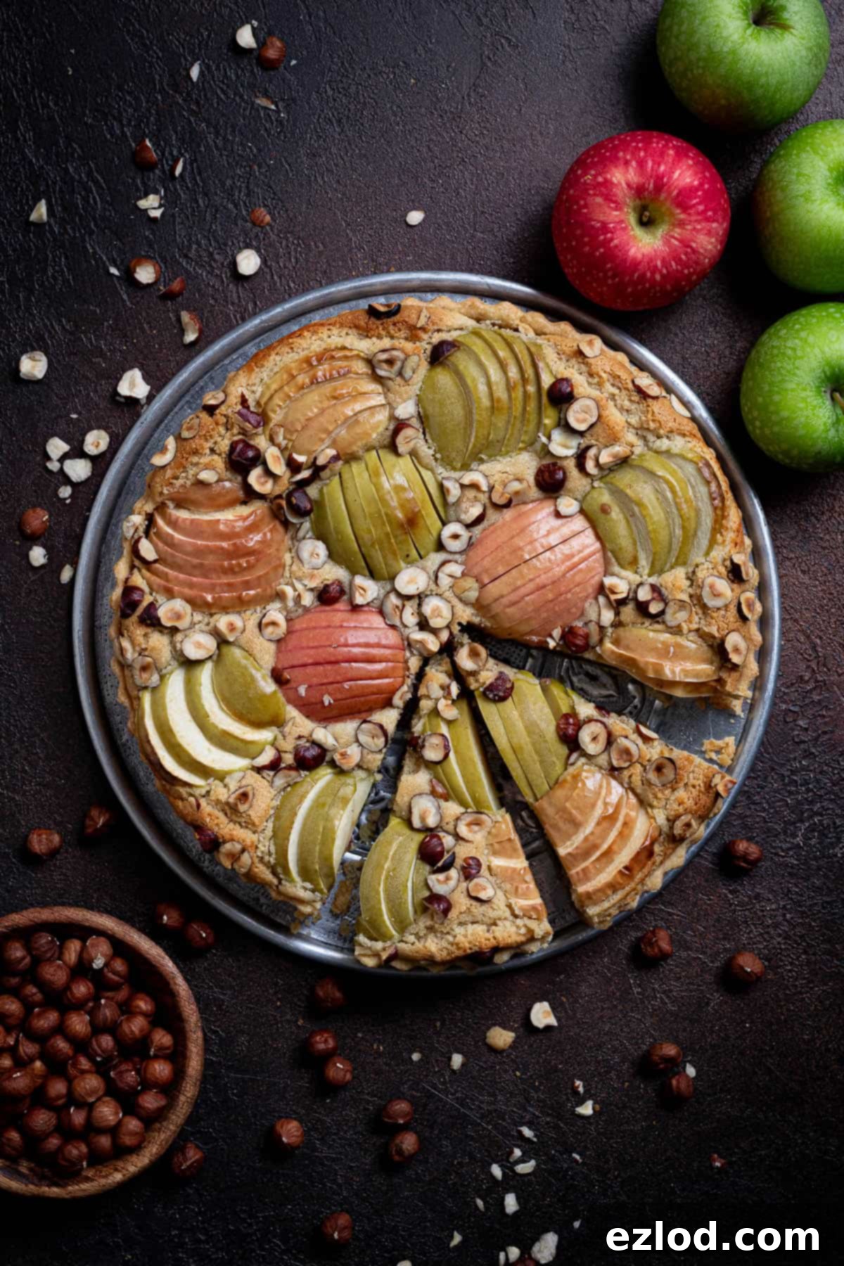 Elegant Vegan Apple Hazelnut Frangipane Tart 18 The stunning sliced vegan apple hazelnut frangipane tart resting on a dark surface, surrounded by fresh apples and a bowl of hazelnuts, inviting you to indulge.