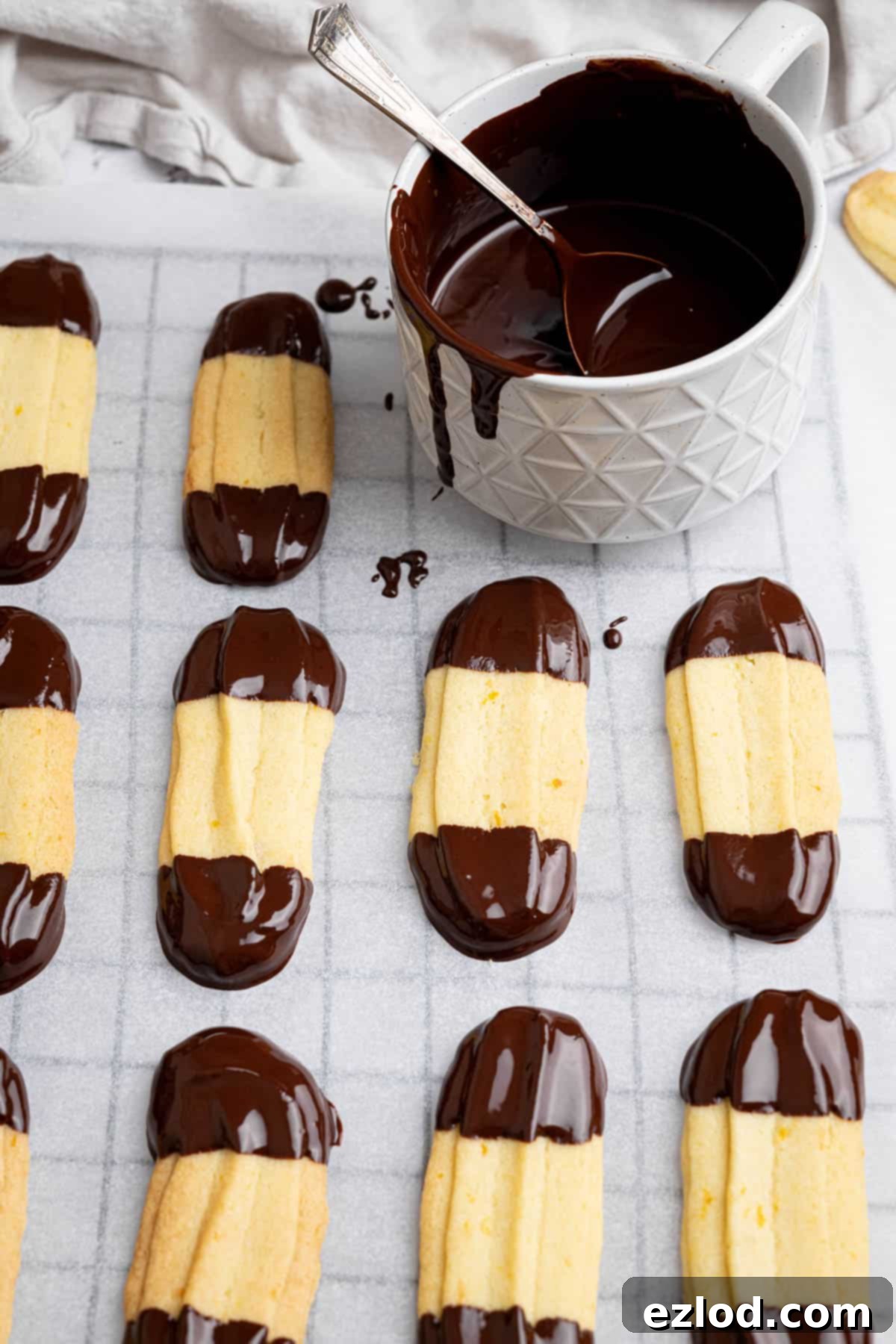 Vegan Chocolate Orange Viennese Fingers 11 Close up of chocolate orange Viennese fingers on a sheet of baking paper with a mug of melted chocolate.