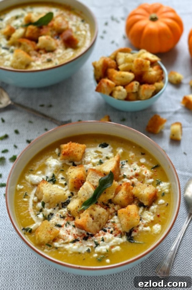 Velvet Pumpkin Soup with Feta, Garlic Croutons, and Fried Sage 10 Overhead view of gently spiced roast pumpkin soup, topped with golden garlic croutons, white feta cheese crumbles, and green fried sage leaves in a white bowl.