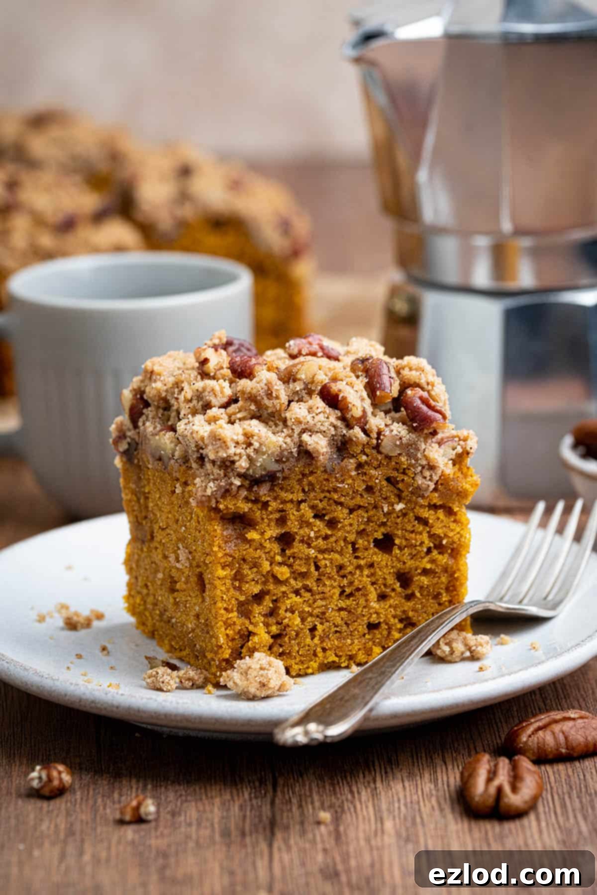 Spiced Vegan Pumpkin Streusel 2 A square of vegan pumpkin crumb cake on a white plate with a fork and a pot of coffee in the background. The cake is moist, rich, and features a crunchy pecan streusel topping, ideal for autumn.
