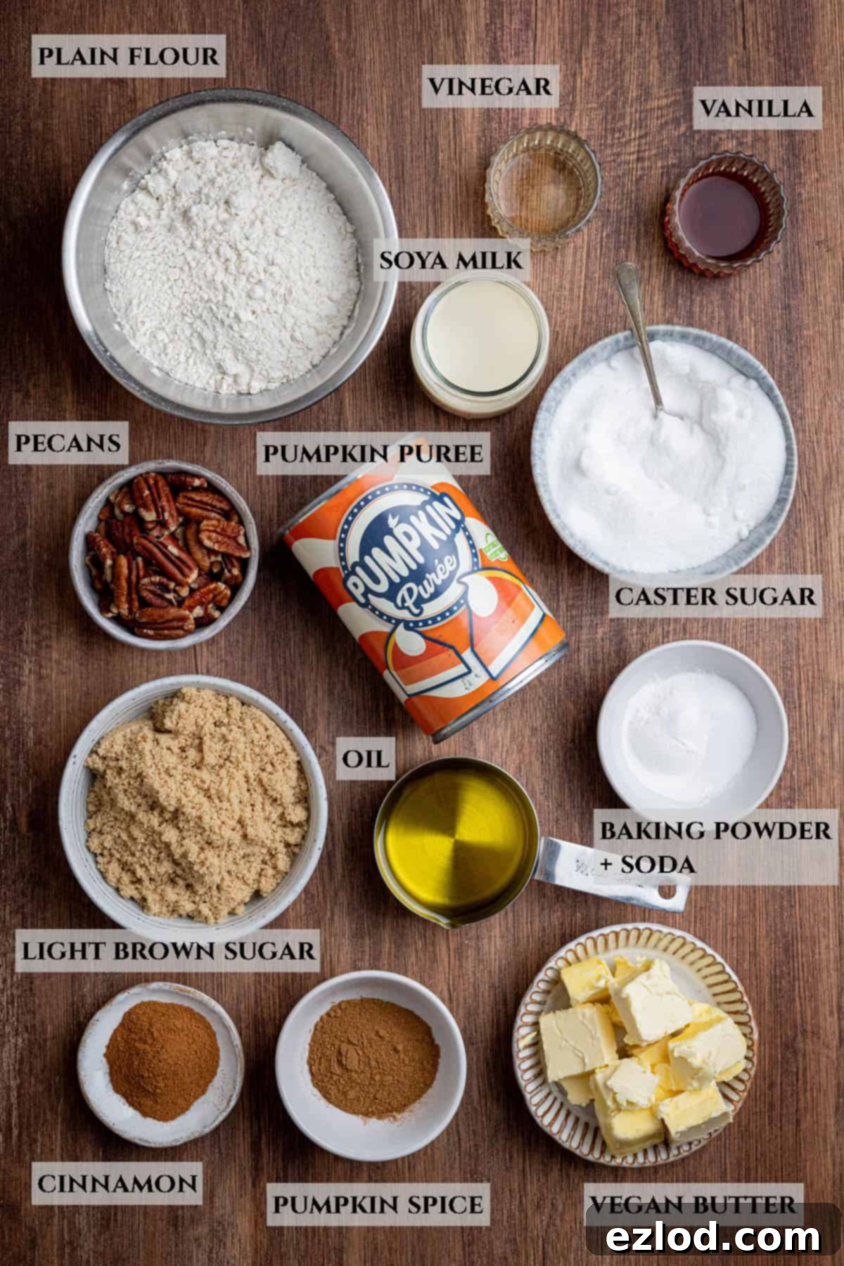 Spiced Vegan Pumpkin Streusel 3 A labelled photo of all the ingredients needed to make a vegan pumpkin crumb cake, laid out in bowls on a white surface, including plain flour, light and caster sugars, pumpkin spice, baking powder, baking soda, salt, pumpkin puree, neutral oil, non-dairy milk, vinegar, vanilla extract, vegan block butter, and chopped pecans.