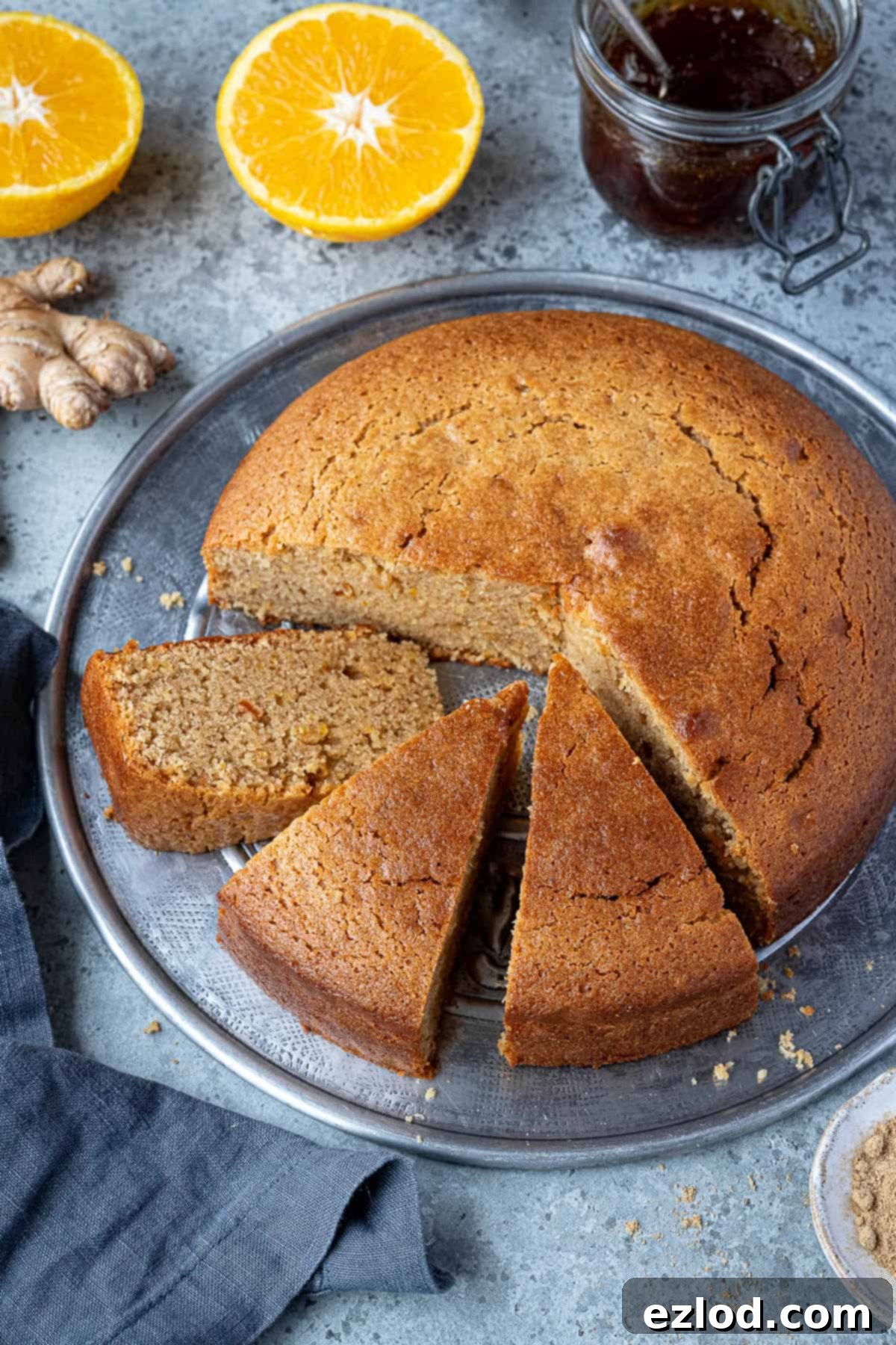 Sun-Kissed Vegan Ginger Cake with Tangy Marmalade 9 A beautifully arranged photograph featuring a sliced vegan marmalade and ginger cake on a metal plate, surrounded by fresh oranges, ginger root, and a jar of marmalade, creating a visually appealing scene.