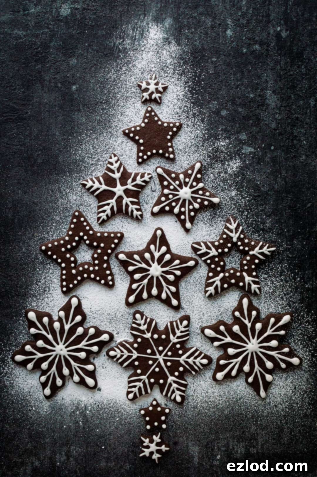 Vegan Chocolate Gingerbread Cookies 6 Iced chocolate gingerbread snowflake biscuits arranged into a Christmas tree shape with icing sugar on a dark background.