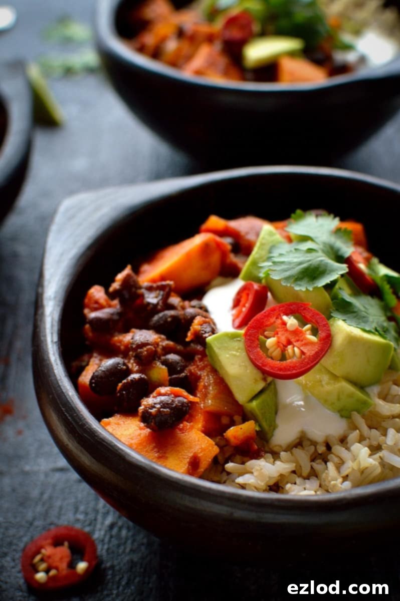 Hearty Vegan Sweet Potato Black Bean Chili 4 A close-up shot of creamy sweet potato and black bean chilli in a rustic black bowl, artfully served with fluffy brown rice, a dollop of cooling sour cream, fresh avocado slices, a sprinkle of vibrant red chilli, and fragrant coriander leaves. This image highlights the texture and inviting presentation of a delicious vegan meal.