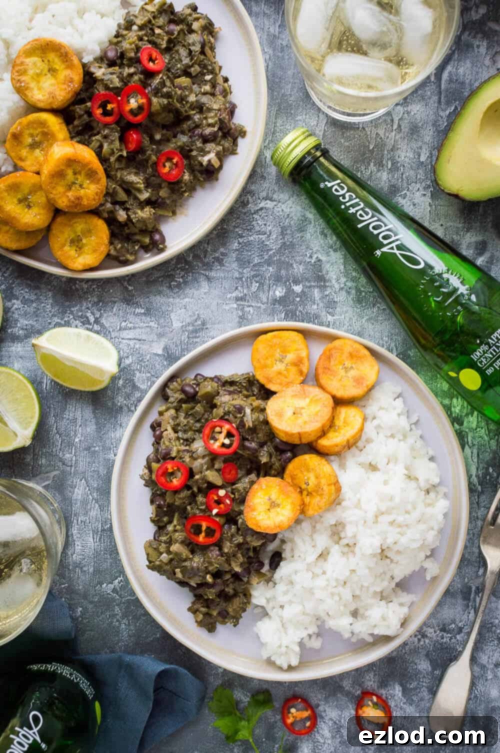 Aromatic Caribbean Medley with Coconut Rice and Sweet Plantain 2 Two plates of vegan spiced black beans and callaloo with coconut rice and fried plantain on a grey background with lime, avocado and Appletiser.