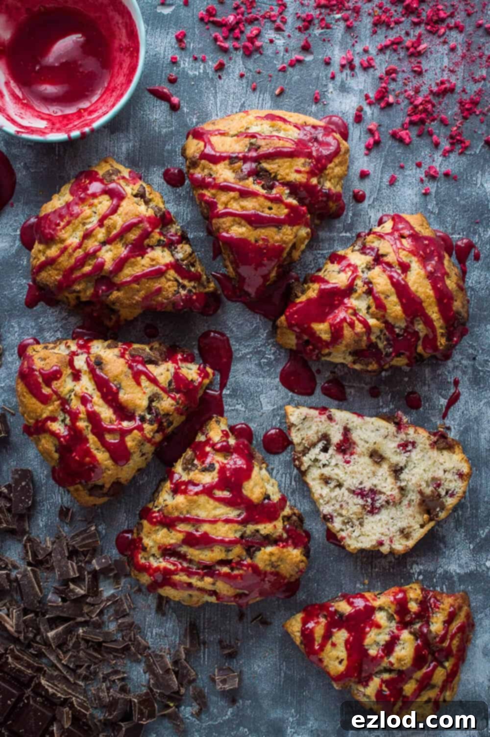 Decadent Vegan Chocolate Raspberry Scones 5 six vegan chocolate raspberry scones with raspberry glaze on a grey background, one sliced in half.