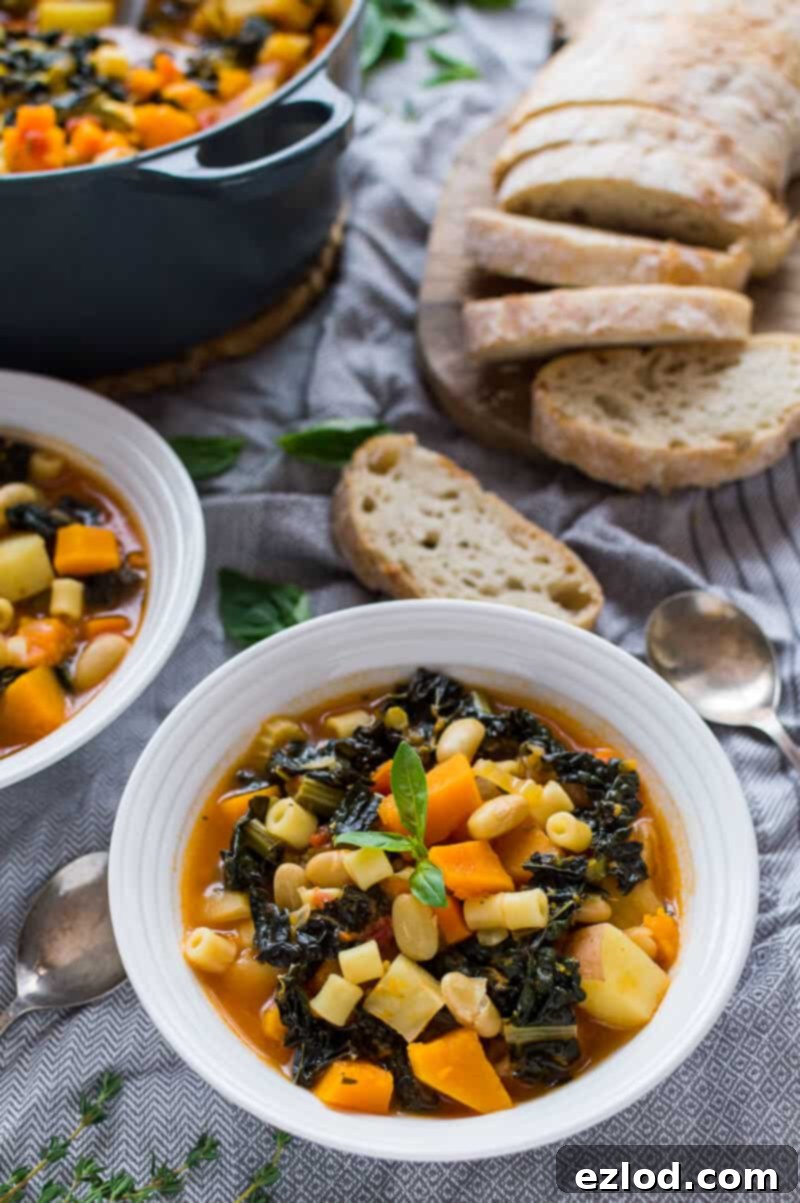 Cozy Vegan Fall Minestrone 8 A beautifully presented bowl of Autumn minestrone soup, garnished with fresh basil, on a grey tablecloth, with crusty ciabatta bread and a large pot of soup in the soft-focus background, ready to be enjoyed.