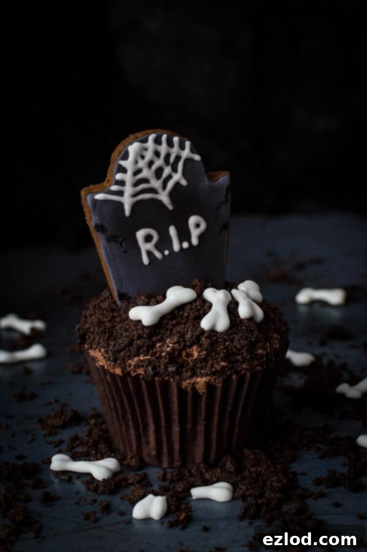 Plant-Based Graveyard Halloween Treats 17 A single vegan tombstone cupcake on a dark background surrounded by Oreo soil and vegan royal icing bones.