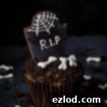 Plant-Based Graveyard Halloween Treats 18 A single vegan tombstone cupcake on a dark background surrounded by Oreo soil and vegan royal icing bones.