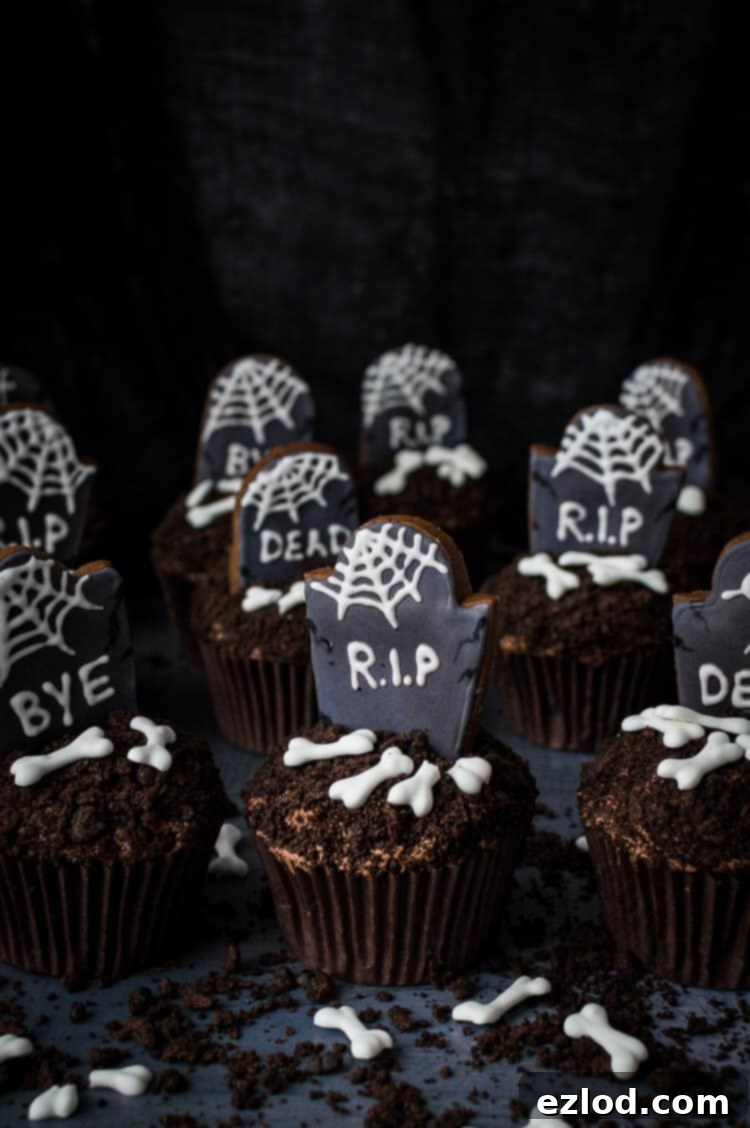 Plant-Based Graveyard Halloween Treats 3 Vegan chocolate cupcakes topped with crushed Oreos, gingerbread tombstones and vegan royal icing bones on a dark background.