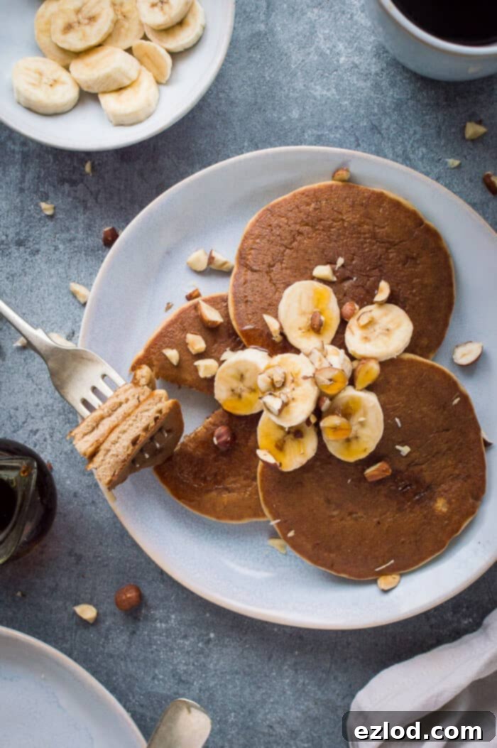 Pillowy Plant-Based Banana Pancakes 6 A plate of three fluffy vegan banana pancakes topped with sliced banana, hazelnuts and maple syrup with a forkful taken out on a grey background.