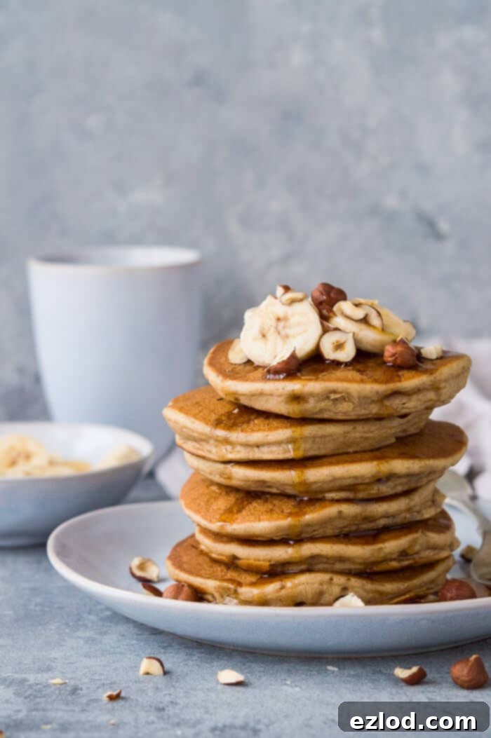 Pillowy Plant-Based Banana Pancakes 7 A stack of fluffy vegan banana pancakes with sliced banana, hazelnuts and maple syrup with a bowl of banana and a cup of coffee.