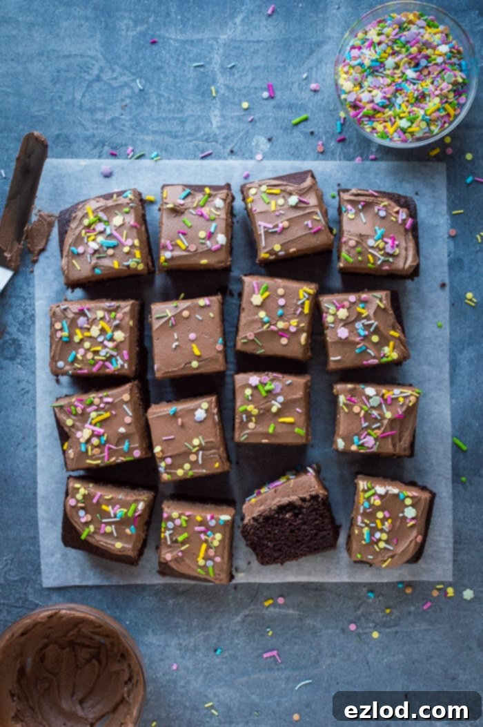 Easy vegan chocolate cake - this simple vegan chocolate sheet cake is quick and easy to make and doesn't use any unusual or hard to find ingredients. It is so moist and rich you would never guess that it is free from eggs and dairy! An appetizing overhead shot of easy vegan chocolate cake, already sliced into perfect squares and arranged on a grey background, accompanied by a bowl of vibrant sprinkles and a bowl of creamy buttercream.