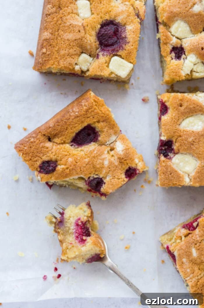 Raspberry, white chocolate and almond sheet cake - this amazing eggless, dairy free and vegan traybake cake is wonderfully soft and moist and full of fresh raspberries and white chocolate chunks. Perfect for snacking, afternoon tea or dessert. #vegan #vegancake #veganbaking #plantbased #dairyfree #eggless #traybake #almondcake Multiple slices of vegan raspberry, white chocolate and almond sheet cake arranged on white baking parchment, with one slice having a forkful removed, highlighting its soft crumb and fruit inclusions. This plant-based dessert is perfect for snacking or an impressive afternoon tea.