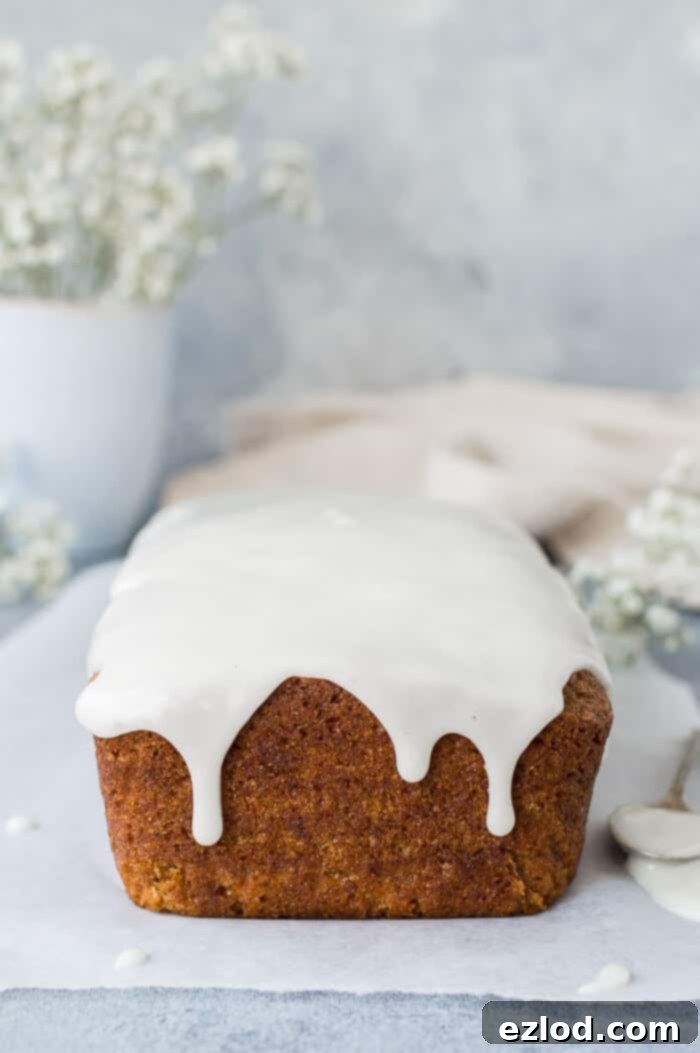 Vegan carrot cake loaf - this is the best vegan carrot cake! Easy to make, moist, sweet and spicy with a zingy lemon glaze; this loaf cake is perfect for snacking on. #vegan #carrotcake #vegancake #veganbaking A full, unsliced vegan carrot cake loaf, elegantly drizzled with lemon glaze, resting on baking parchment. Set against a serene grey background, accompanied by delicate white flowers, evoking a sense of calm and natural beauty.